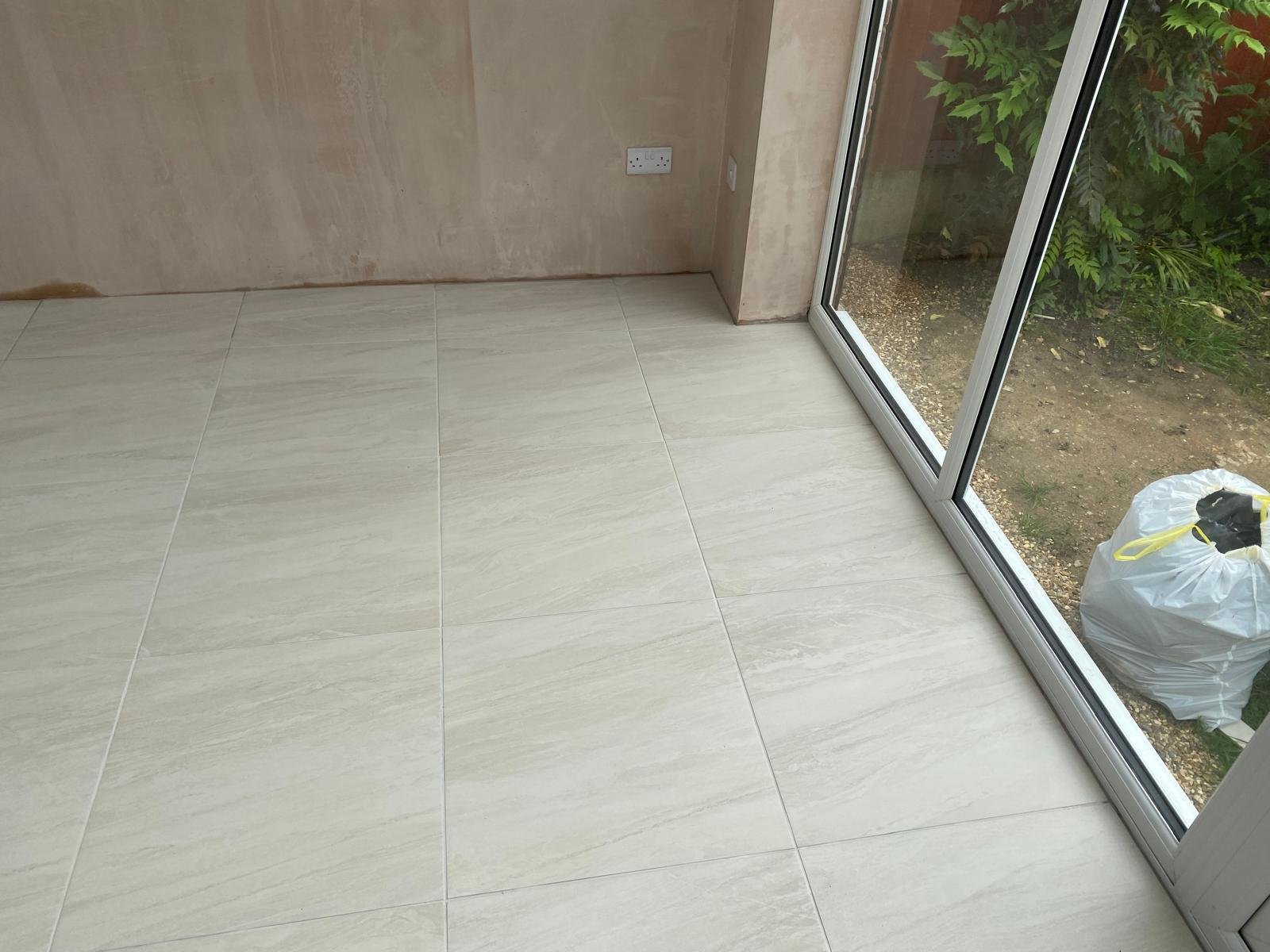Empty room with beige tiled floor, partially plastered wall, electrical outlet, and sliding glass door leading outside to a garden area with a white trash bag outside.