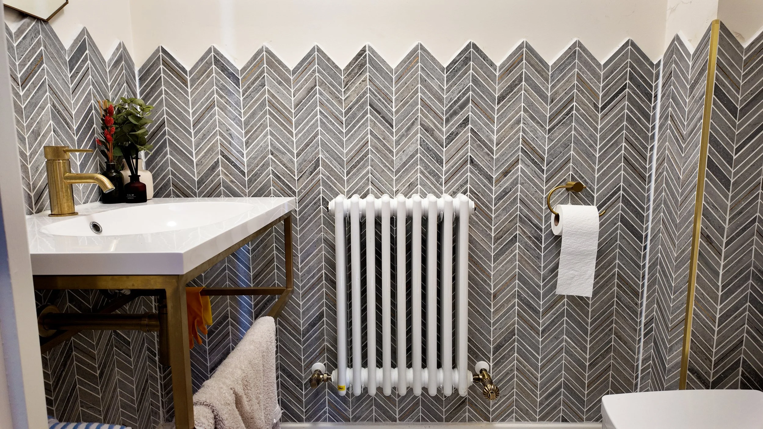 Bathroom with a black, gray, and white herringbone tile wall, a white sink with gold fixtures, a beige towel hanging below, a white radiator, and a toilet paper holder with a roll of toilet paper.