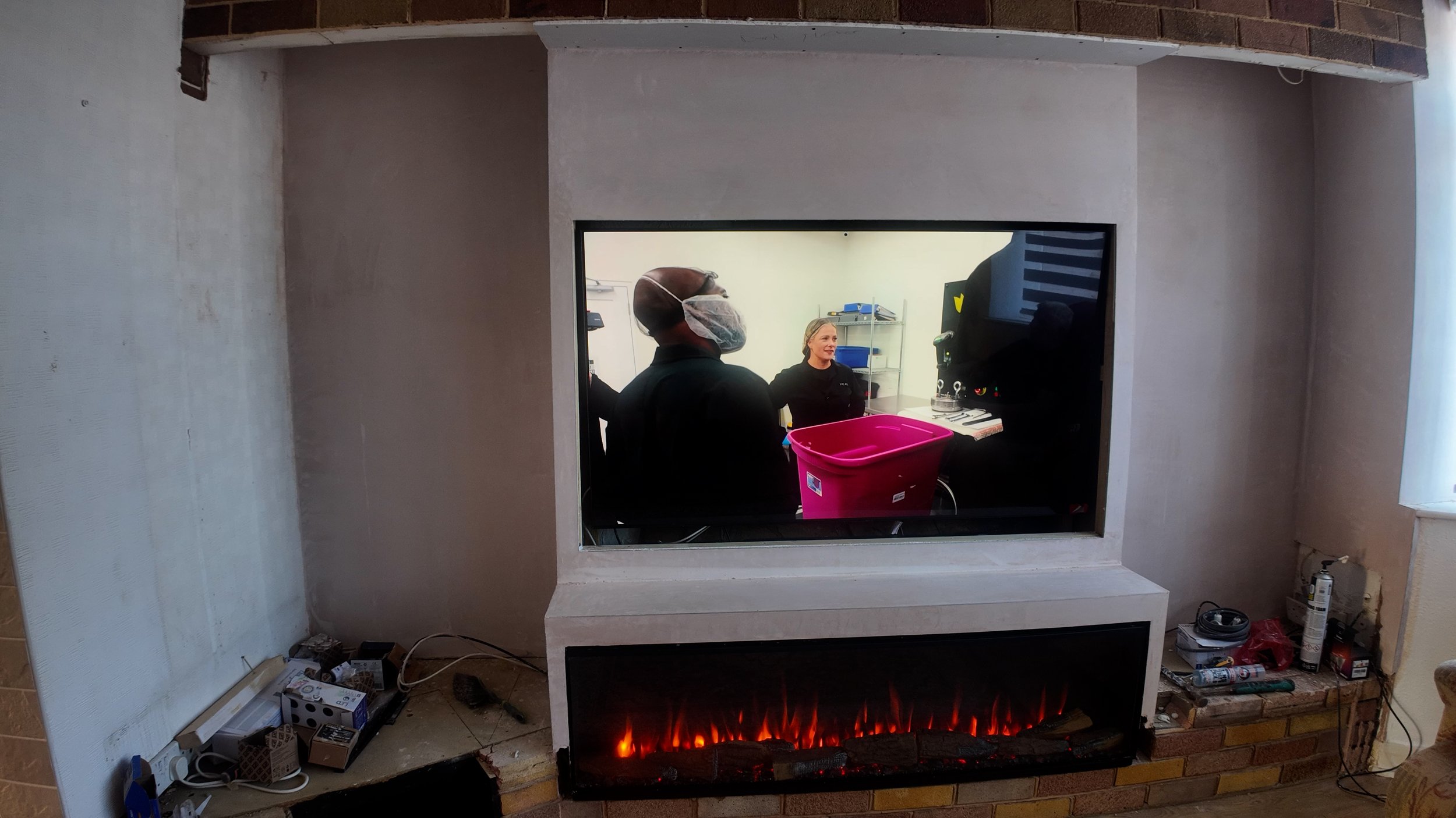 View of a fireplace with a viewing window showing a laboratory or medical setting behind it, where two people are talking, one wearing a face mask and cap, and the other smiling, with shelves and equipment in the background.