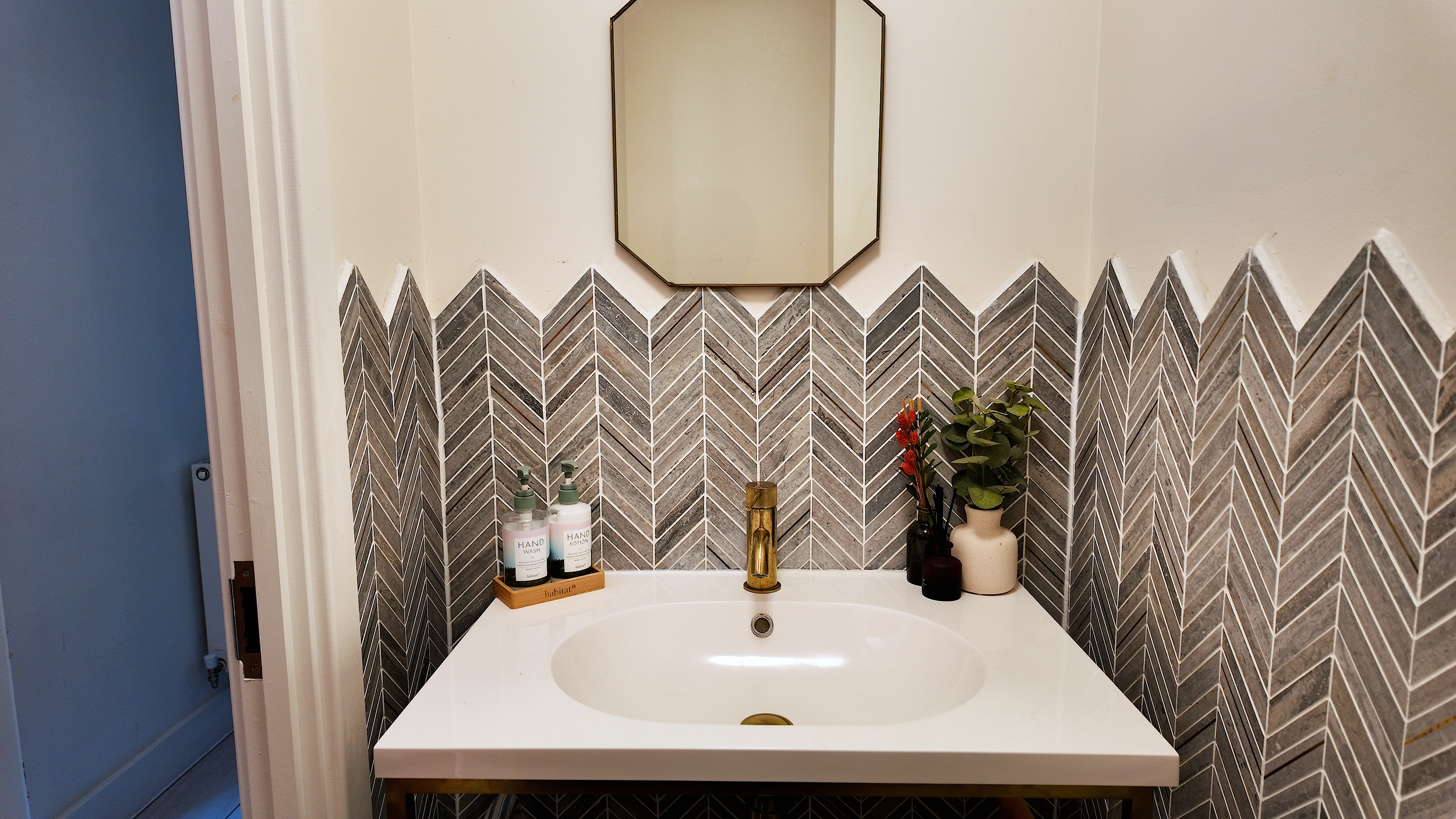 Modern bathroom vanity with a white countertop, gold faucet, decorative vases, hand lotion and soap, and a wall mirror above a wall with gray and white patterned tiles.