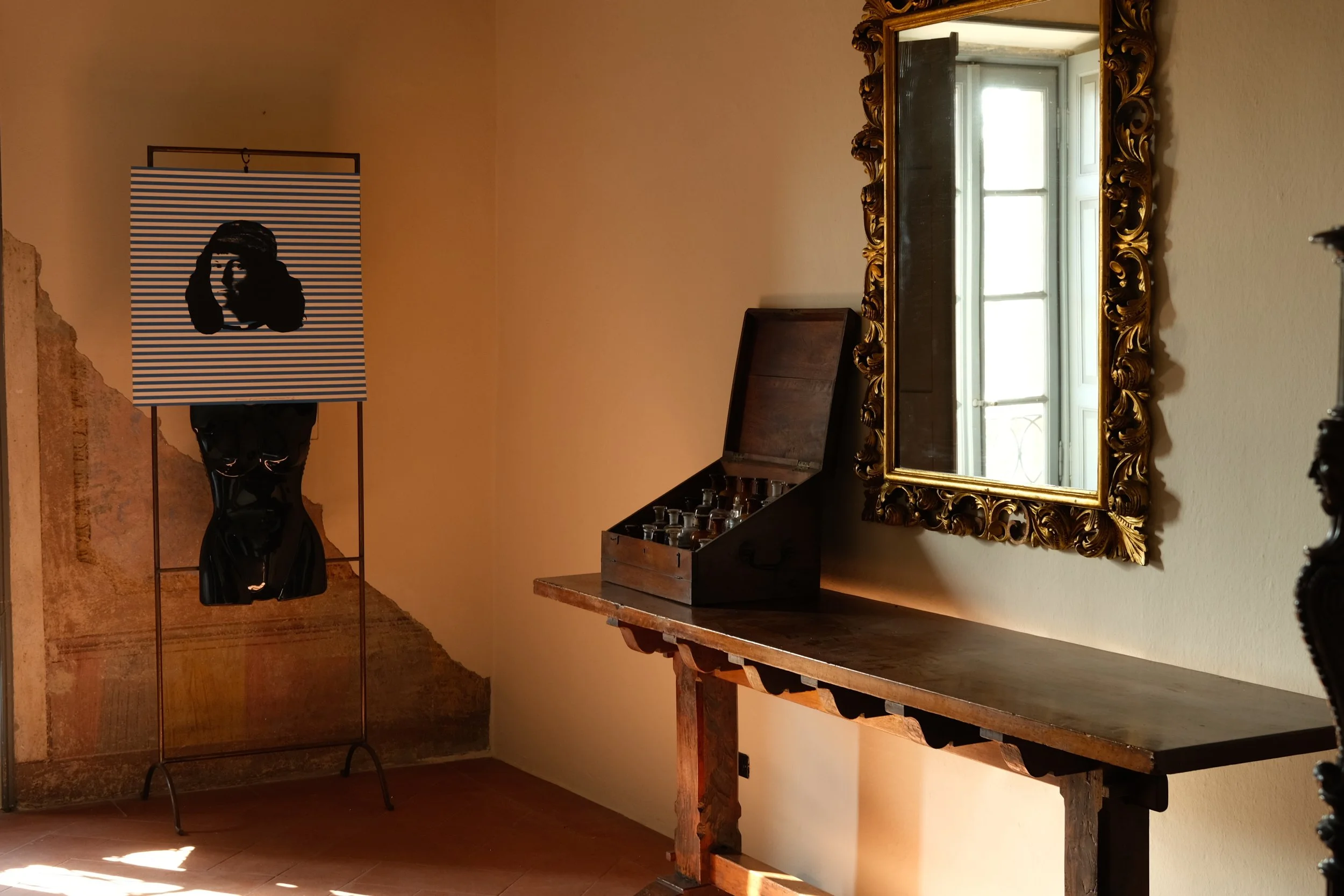 An art display features a bust sculpture with a striped panel on top, a wooden table, an open wooden case with small glass bottles, an ornate gold-framed mirror, and a window reflected in the mirror in a room with beige walls and a partially peeling mural.