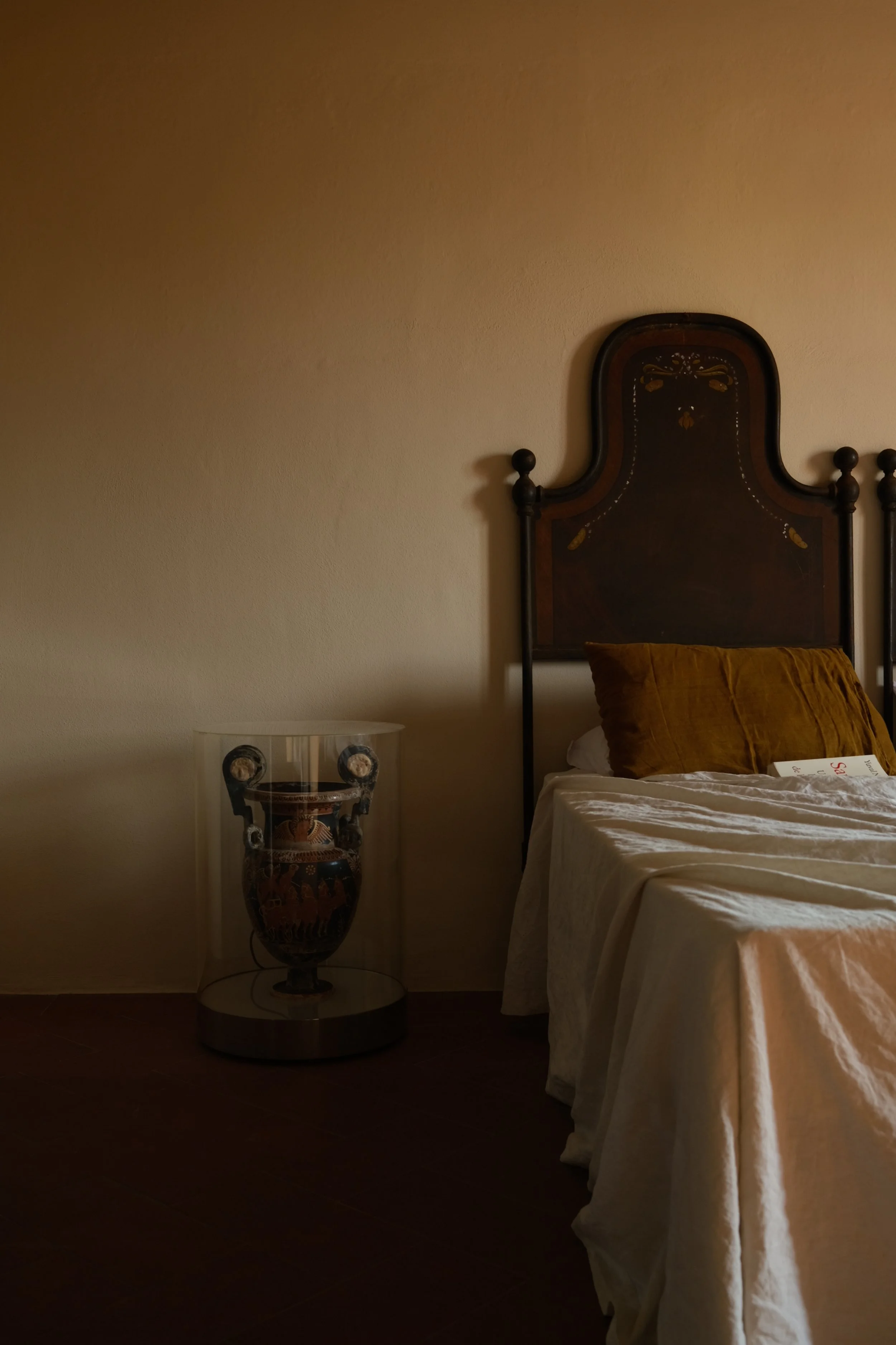A bedroom with a wooden headboard, a bed with a mustard-colored pillow, and a nightstand with an ancient Greek vase inside a glass case.