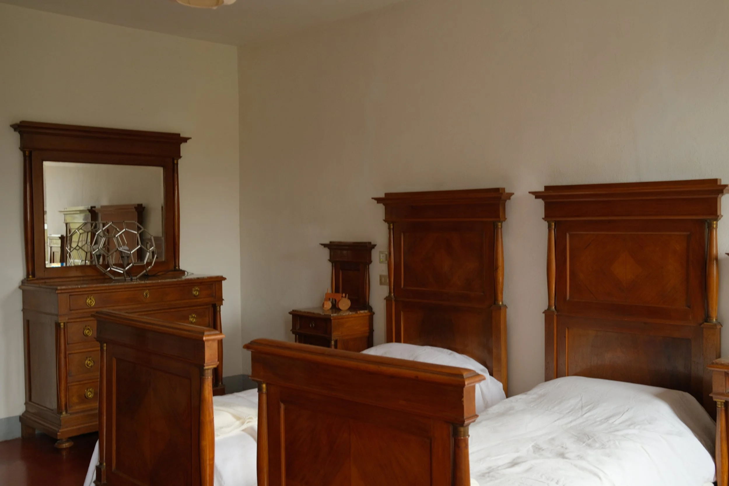 A bedroom with two single beds, each with a tall wooden headboard, white bedding, and a wooden dresser with a mirror behind one of the beds.