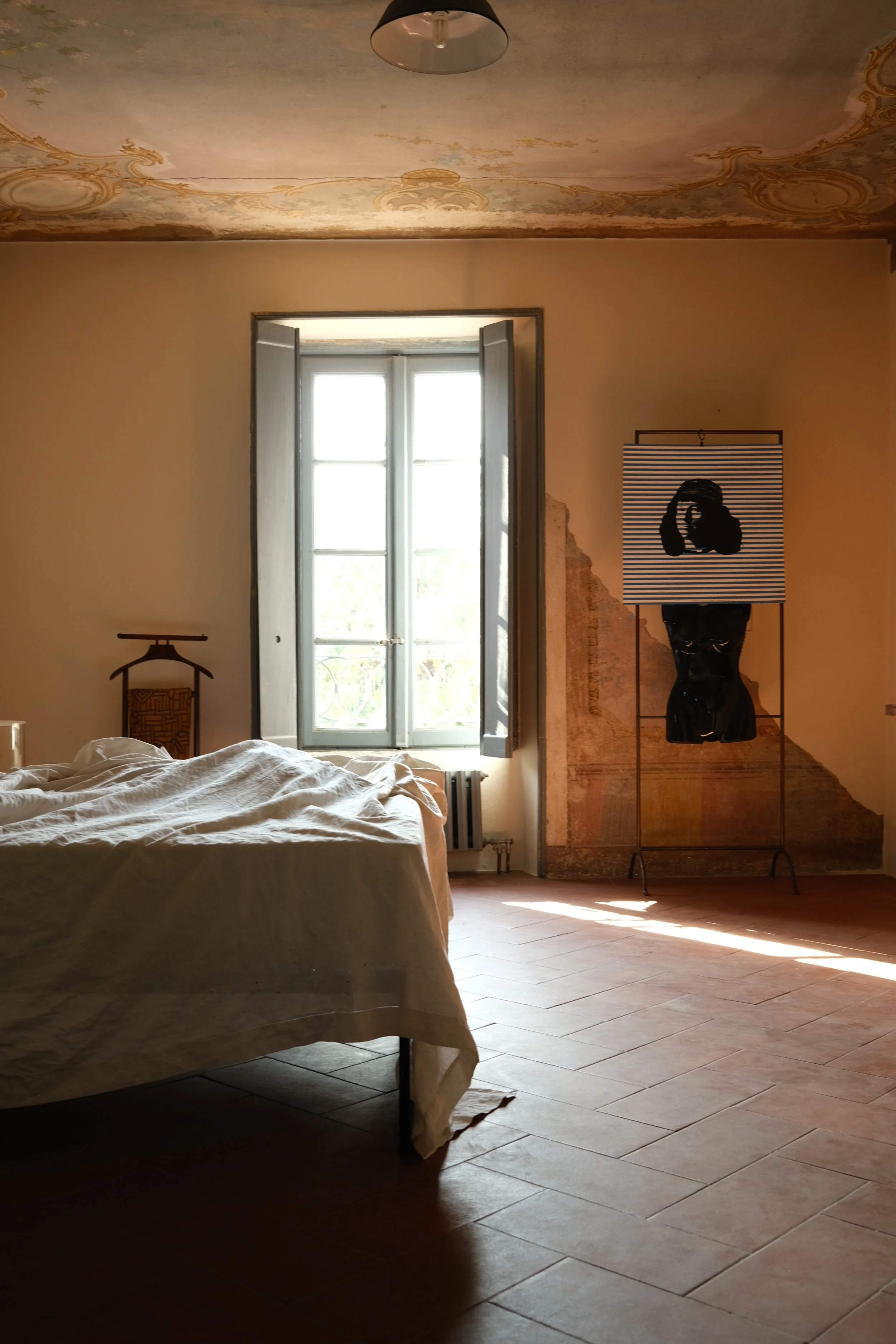 A bedroom with a bed partially visible on the left, an open window letting in natural light, and an art piece with a black figure on a striped background on a stand on the right. The ceiling has decorative painted details, and the floor is tiled.