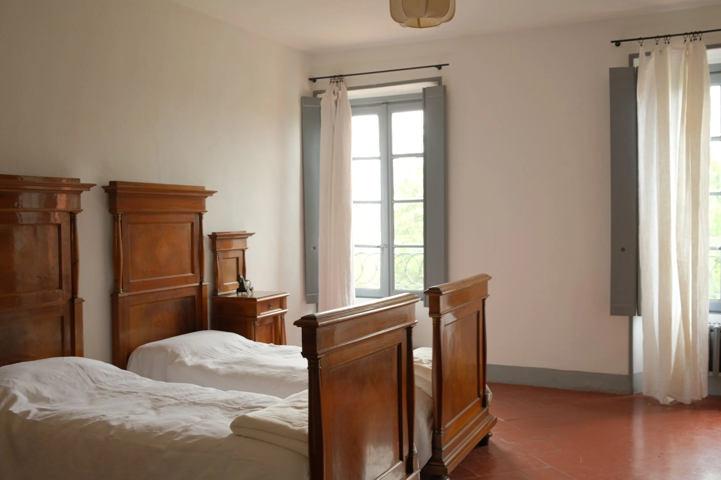 A bedroom with two twin beds with wooden headboards, a small wooden nightstand, two windows with light curtains, and white walls.