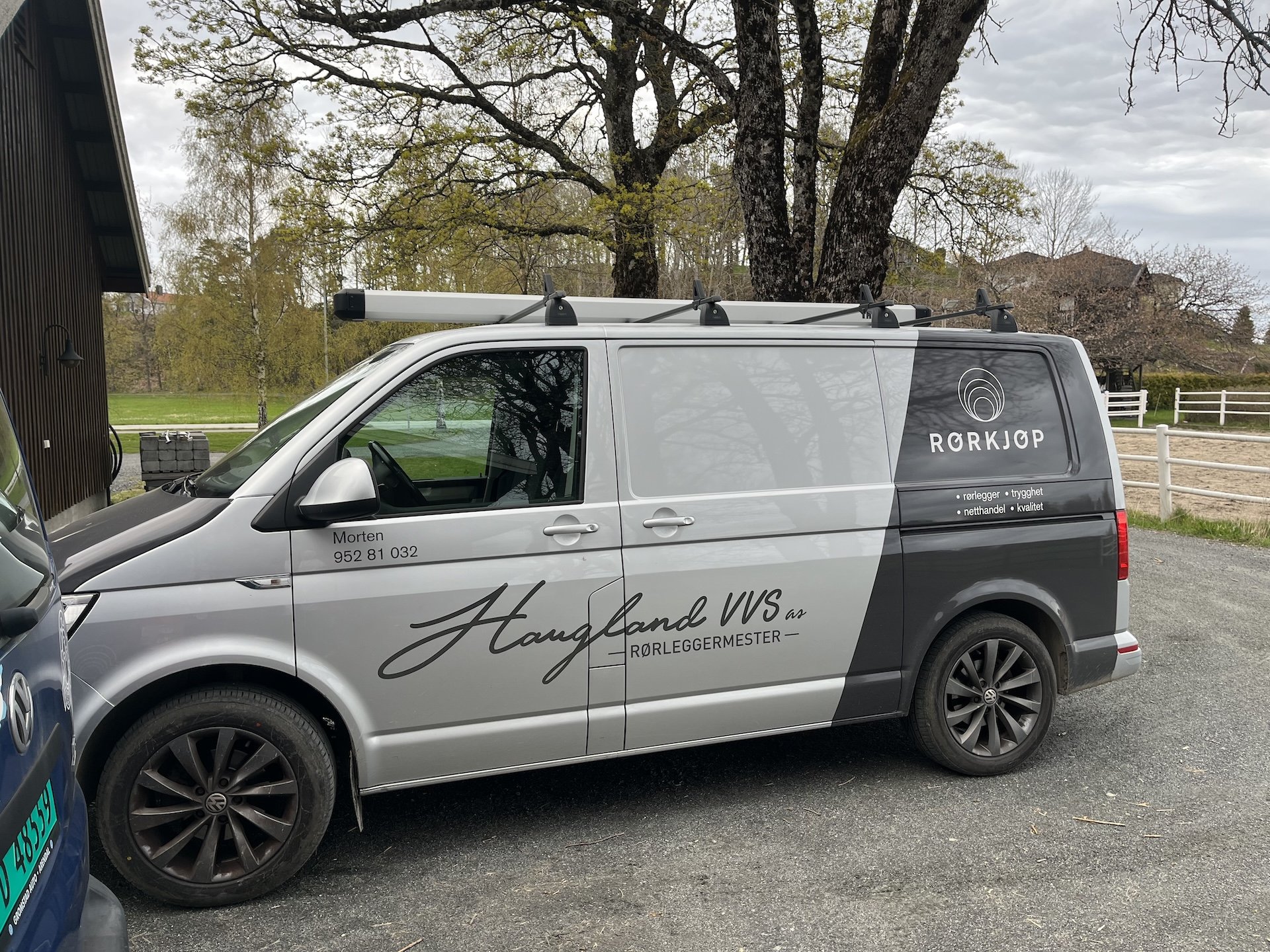 En sølvfarget firmabil med driftsinformasjon og logo, parkert på en vei med trær og bygninger i bakgrunnen.