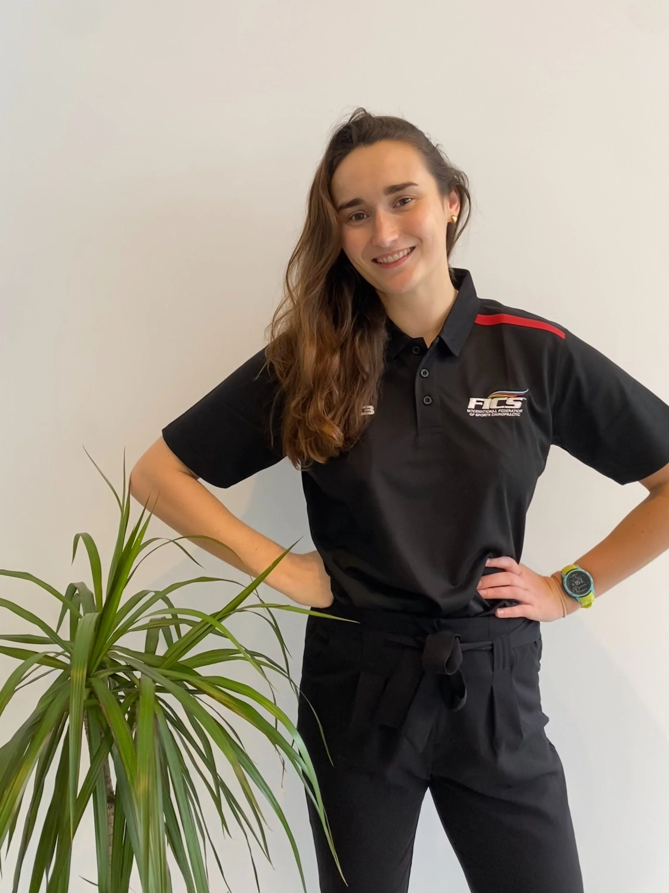 A young woman with long brown hair, wearing a black sports shirt with red and white logo, black pants, and a yellow sports watch, stands confidently with hands on hips beside a green plant against a plain white wall.