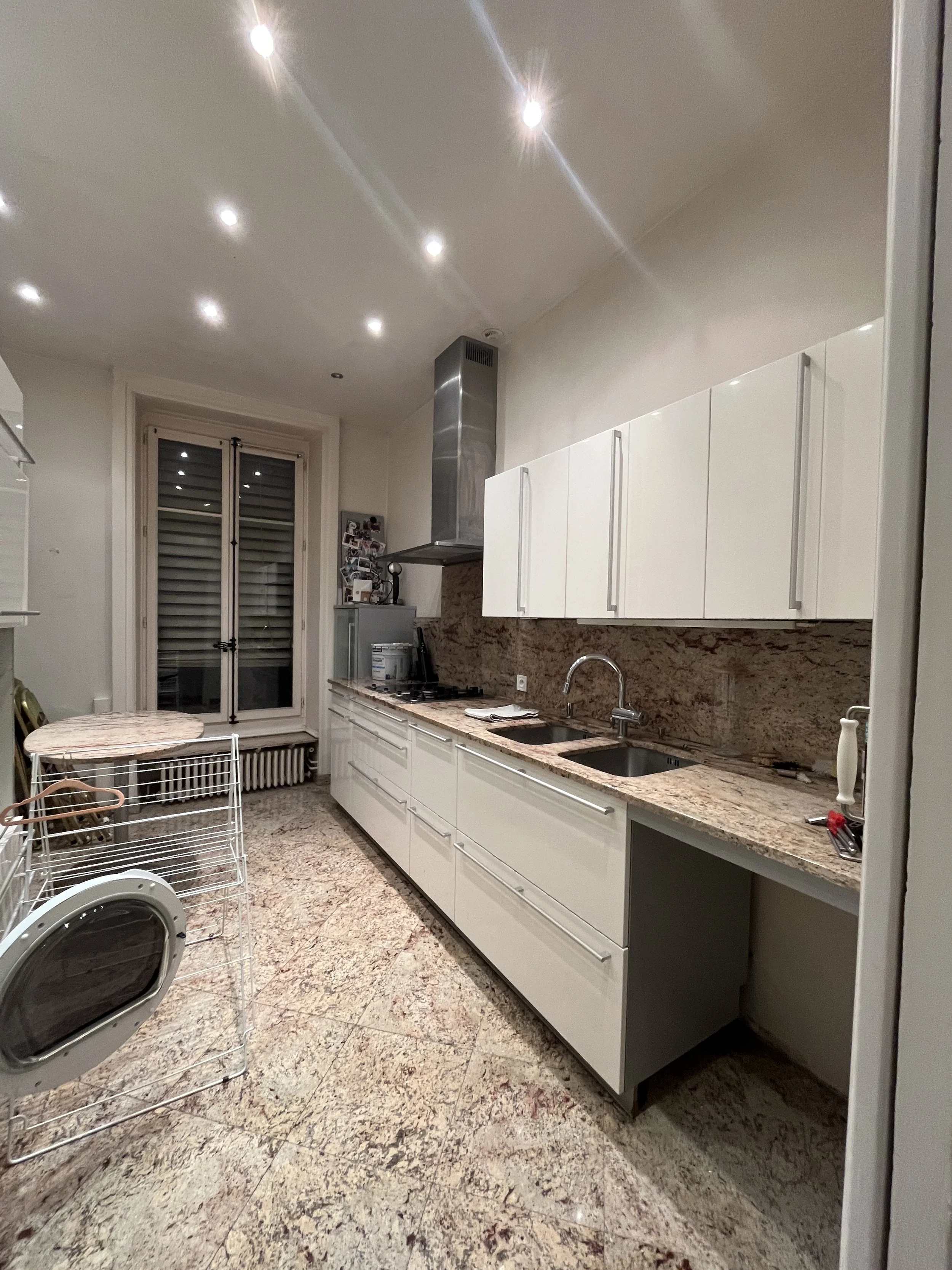 Cuisine moderne avec des armoires blanches, un plan de travail en granit, double évier, planche à découper, et une hotte en acier inoxydable. Il y a un porte-coton et des ustensiles sur le comptoir, une table ronde avec des chaises, une étagère à sèche-linge, et un grand radiateur près d'une porte-fenêtre avec volets roulants.