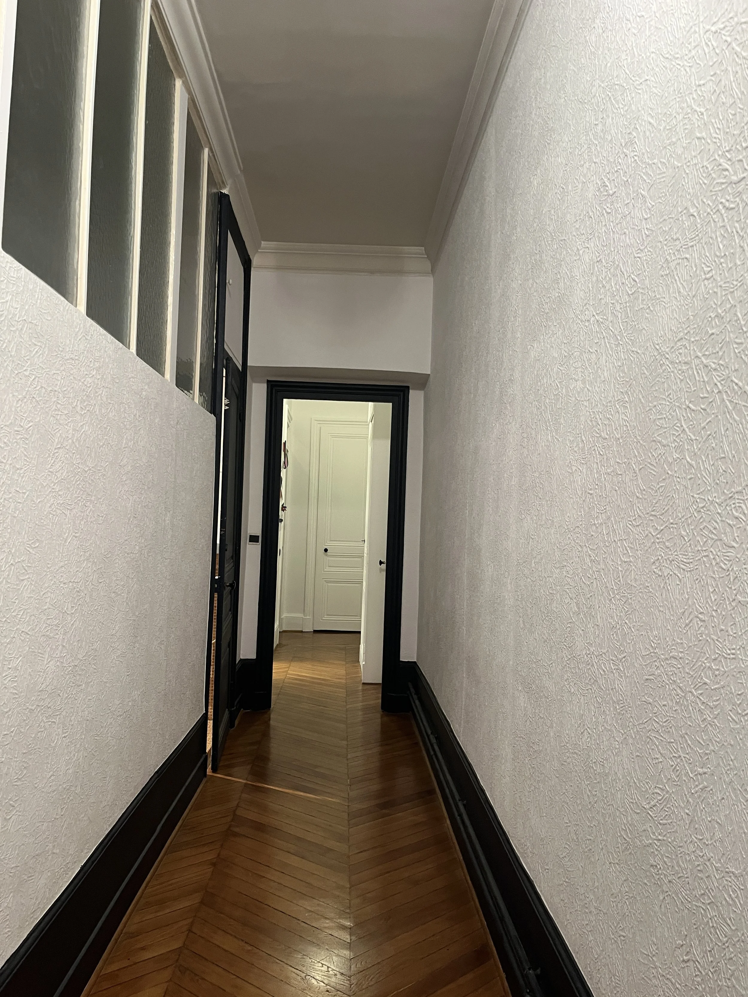 Long couloir intérieur avec murs blancs, sol en bois marqueté, portes noires et fenêtres en haut à gauche.