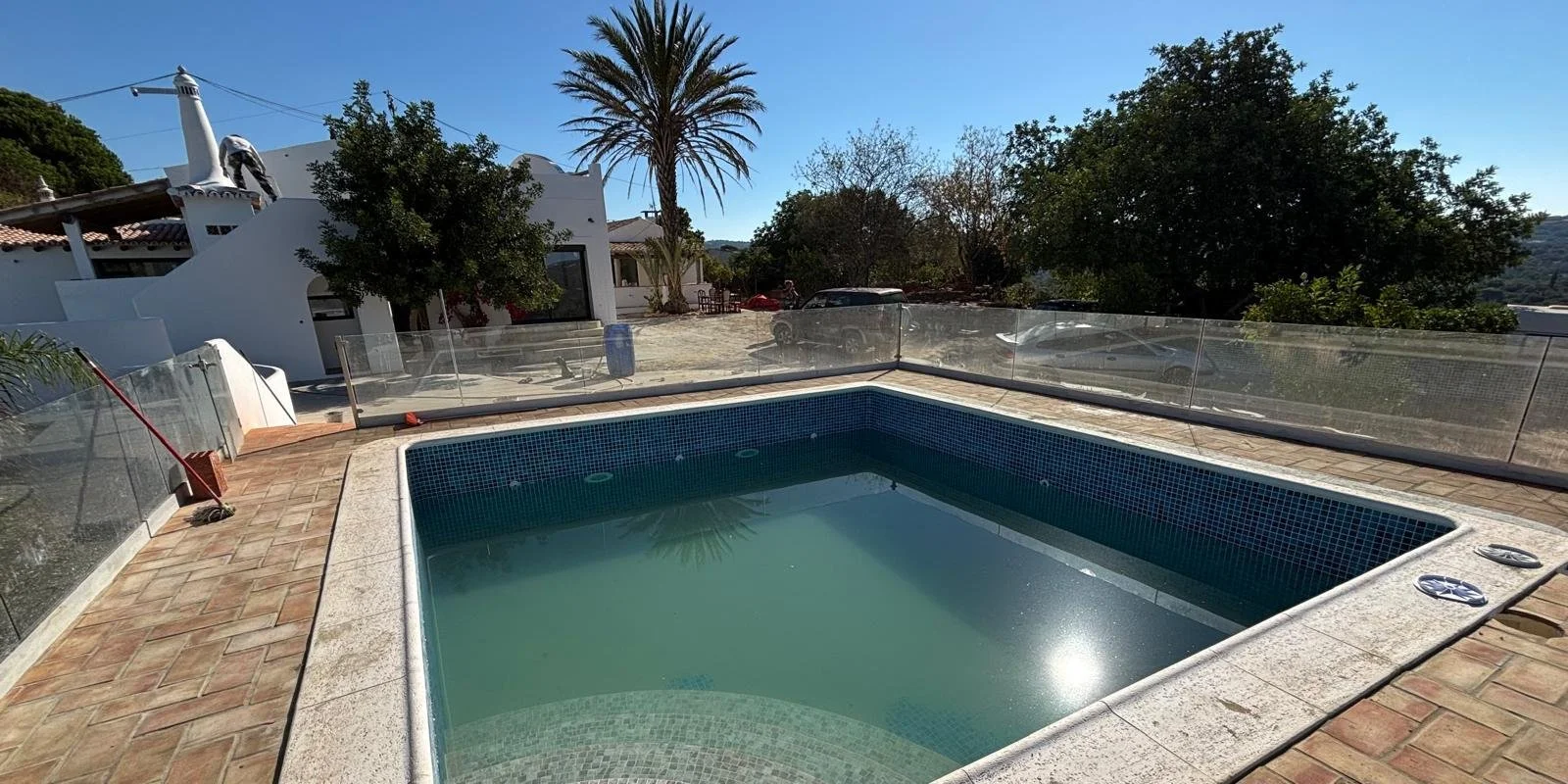 Création piscine entourée d'une terrasse en carrelage, avec une barrière en verre autour. En arrière-plan, une maison blanche avec une palmière et divers arbres, et des voitures garées.