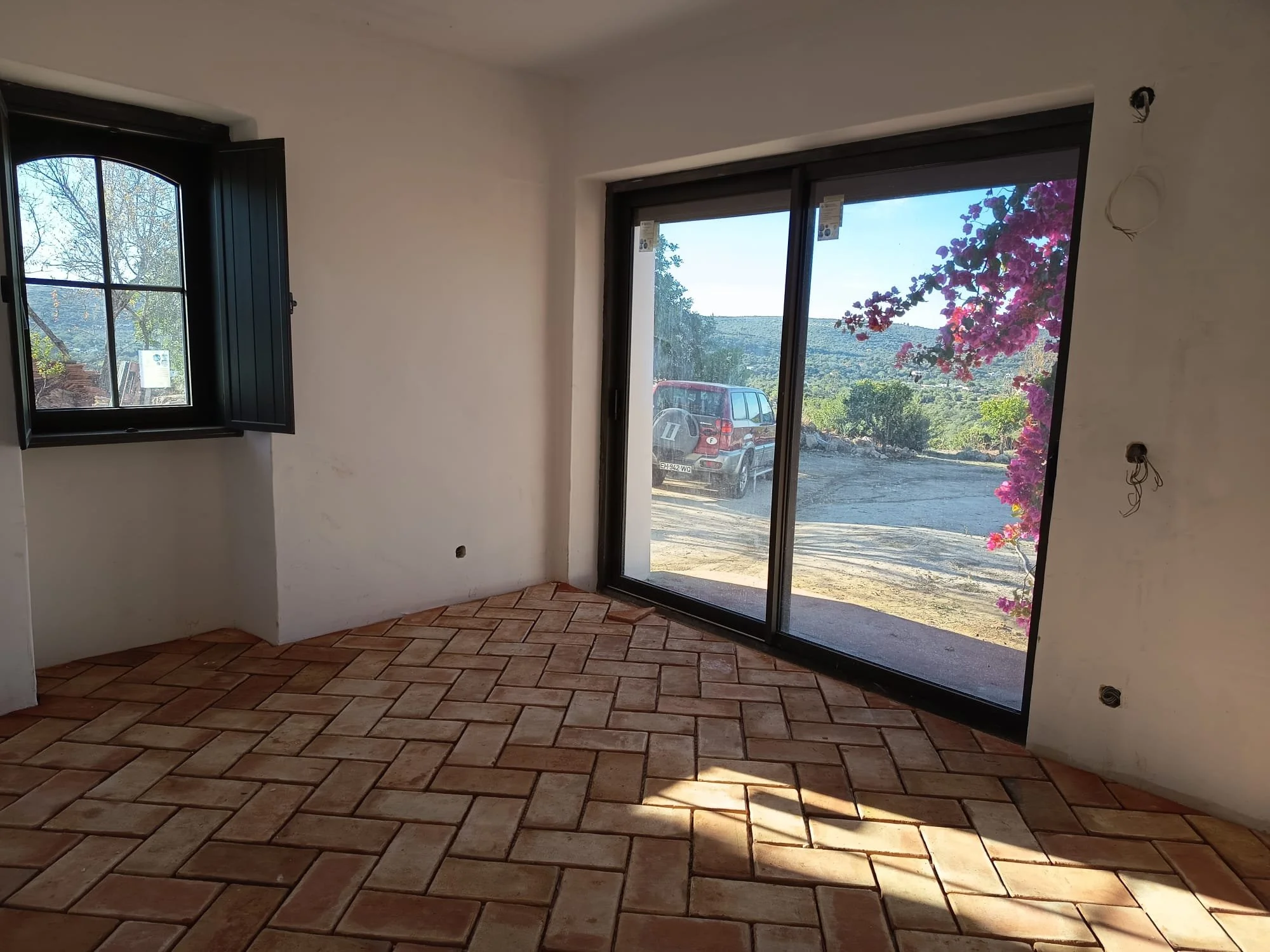 Intérieur d'une pièce en construction avec murs blancs, sol en carreaux de terre cuite, et fenêtres grande baie vitrée offrant une vue sur un paysage rural avec un véhicule et des arbres en fleurs.