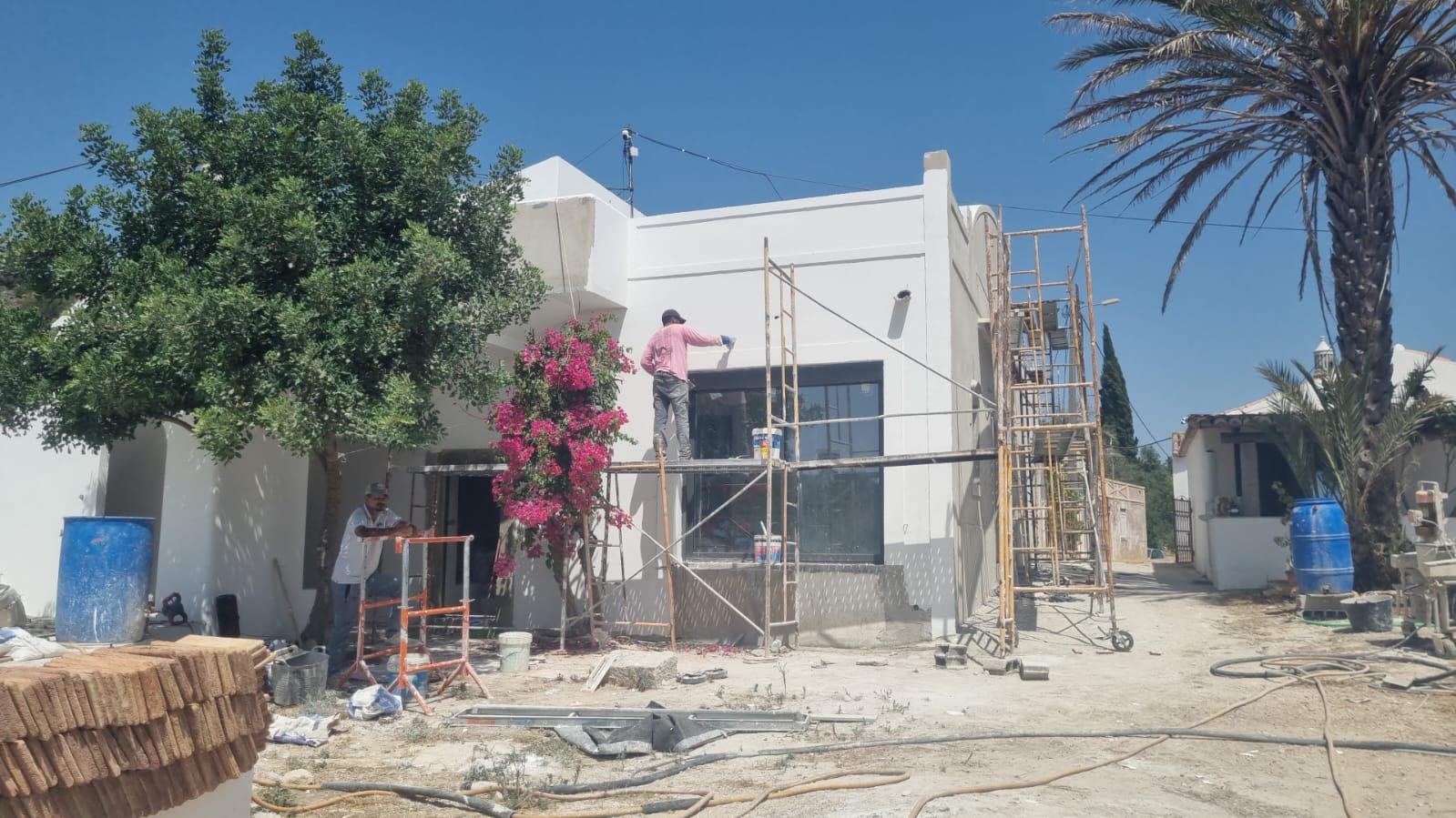 Construction d'une maison blanche au Portugal avec deux ouvriers travaillant sur la façade, échelles, scaffolding, arbres, arbres de bougainvilliers en fleurs roses, ciel bleu clair.