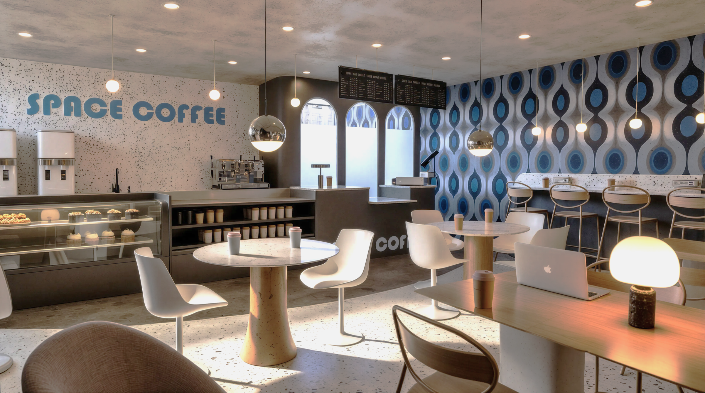 Intérieur d'un café nommé Space Coffee avec murs à motifs bleus, comptoir de service, étagères avec boissons et pâtisseries, tables avec des chaises blanches, lampes suspendues et un ordinateur portable sur une table.