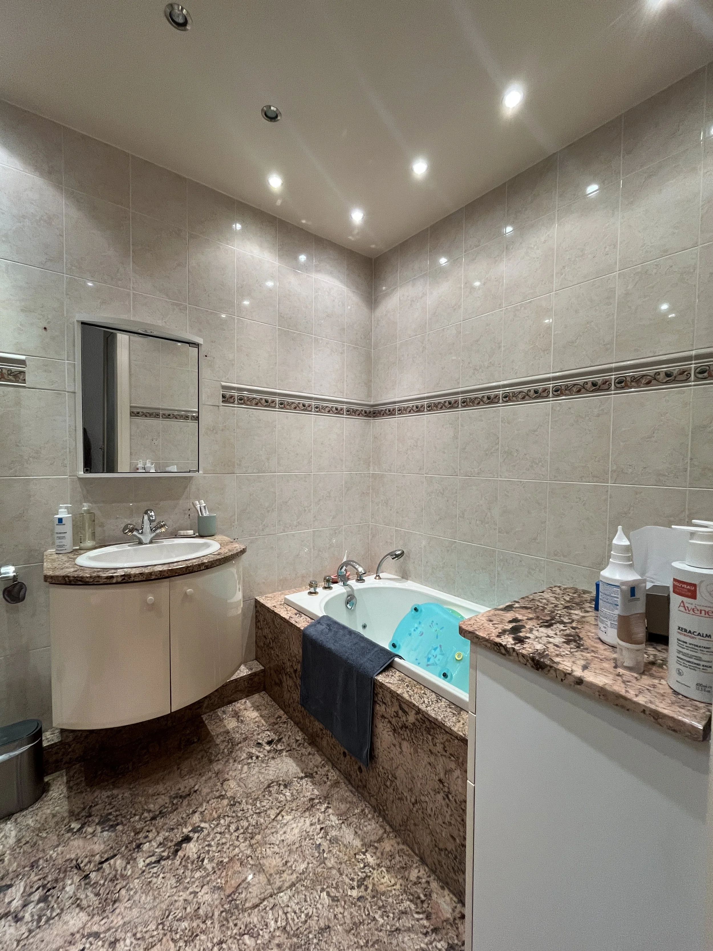 Salle de bain avec baignoire, lavabo, miroir, et revêtement en carrelage beige.