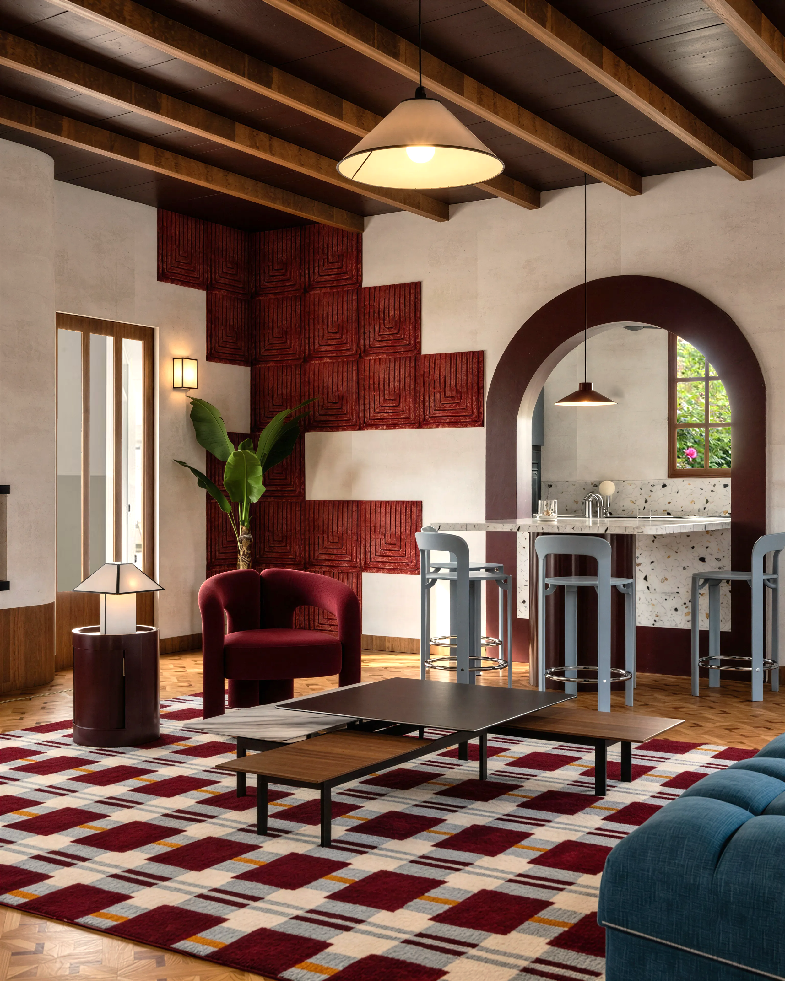 Intérieur moderne comprenant un salon et une cuisine avec un plafond en bois, un tapis géométrique rouge, blanc, gris et jaune, un fauteuil en velours rouge, une table basse en bois et métal, une fresque artistique rouge sur le mur, un mur en plâtre beige, une lampe suspendue beige, un miroir rond dans l'arcade, une grande fenêtre avec vue sur un jardin ensoleillé, et un comptoir de cuisine en marbre avec des tabourets gris.
