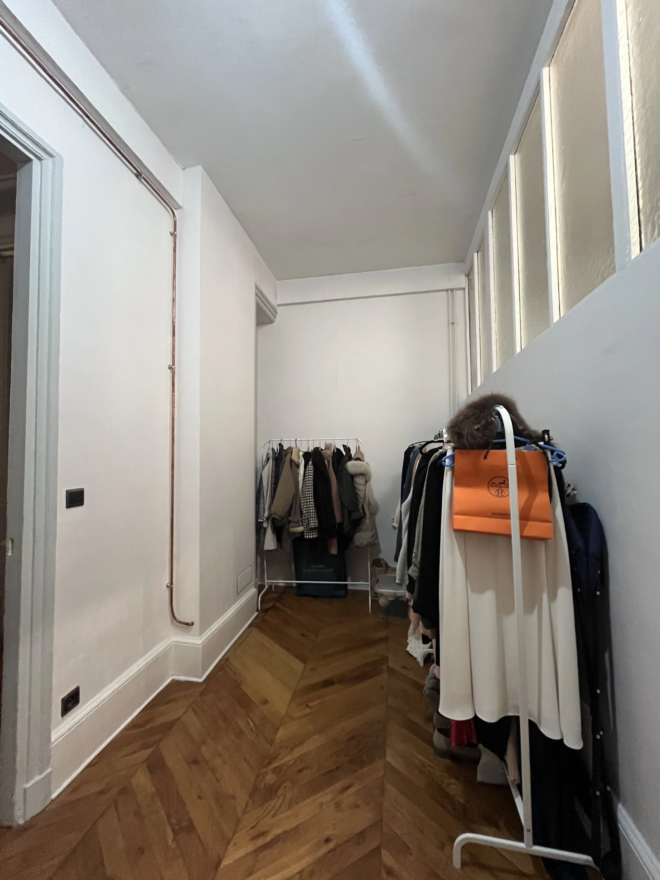 Couloir intérieur avec un mur blanc, un sol en parquet en chevron, un porte-manteaux avec des vêtements et un sac en orange, et une fenêtre en haut à droite de la photo.