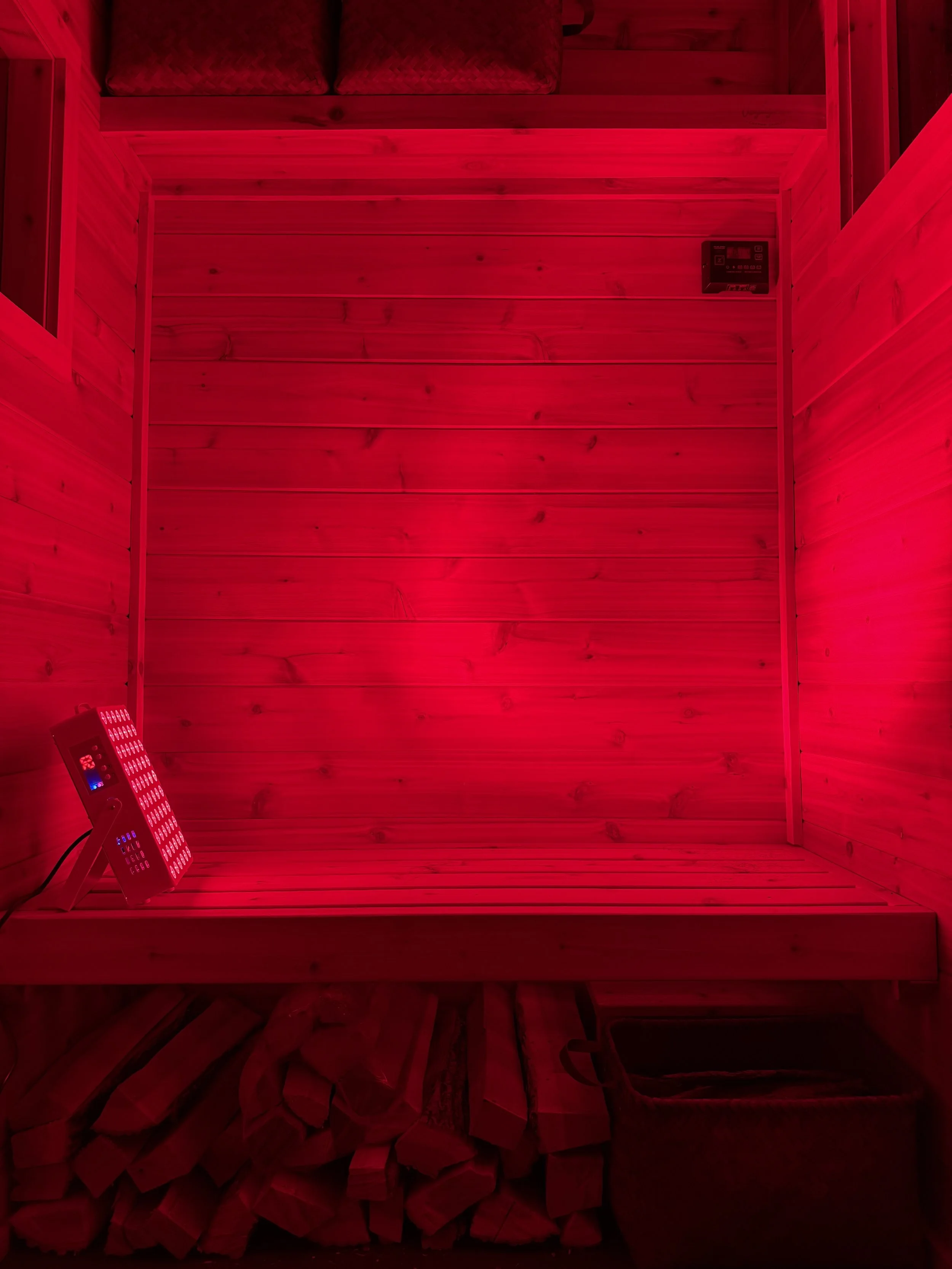 Interior of a wooden sauna with red lighting, a small digital control panel on the wall, and a bench with a bucket underneath.