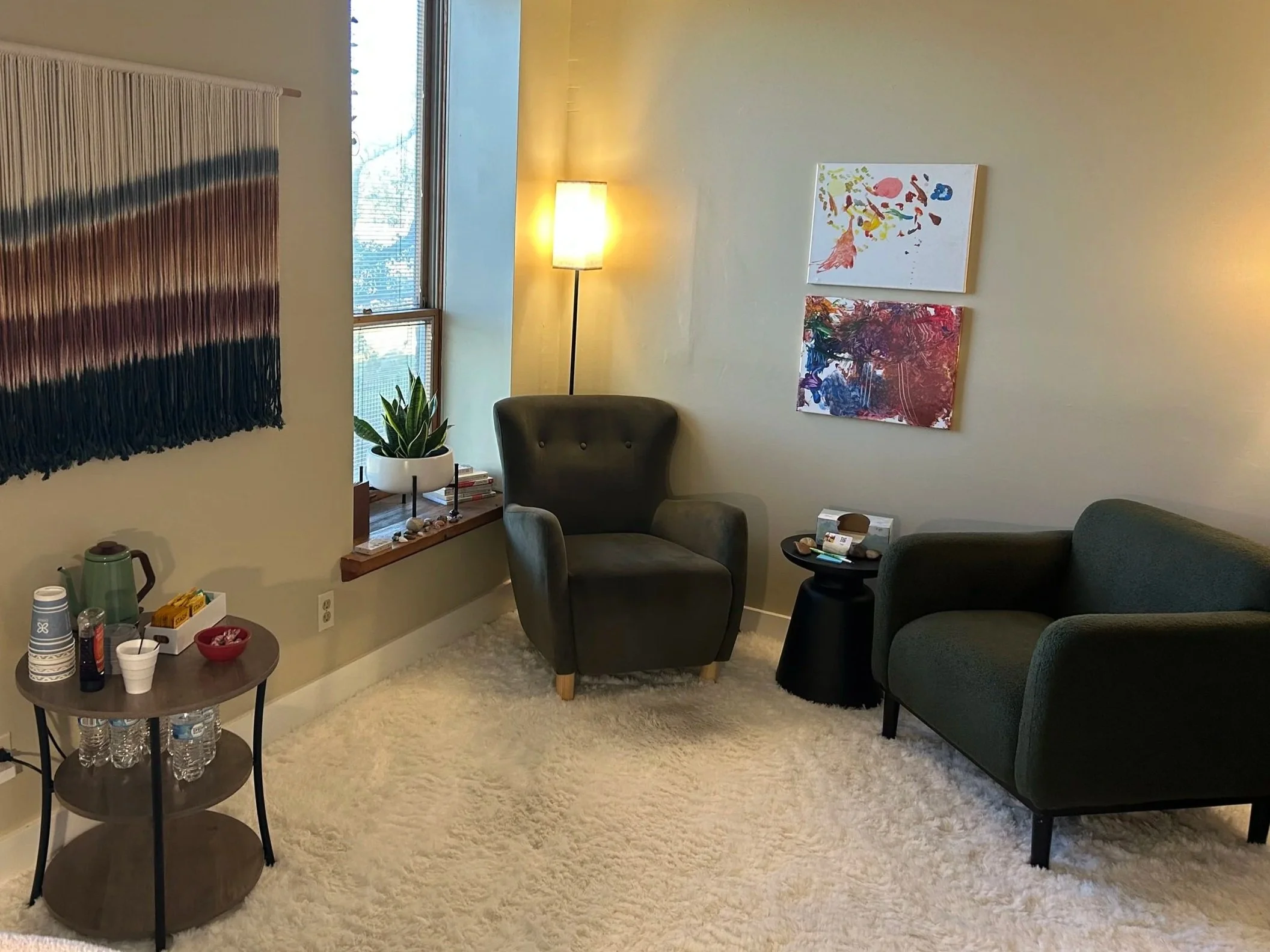 A cozy living room corner with a black armchair, a green sofa, a small black side table, a floor lamp, abstract wall art, a window with a potted plant, and a round wooden table with drinks and snacks.