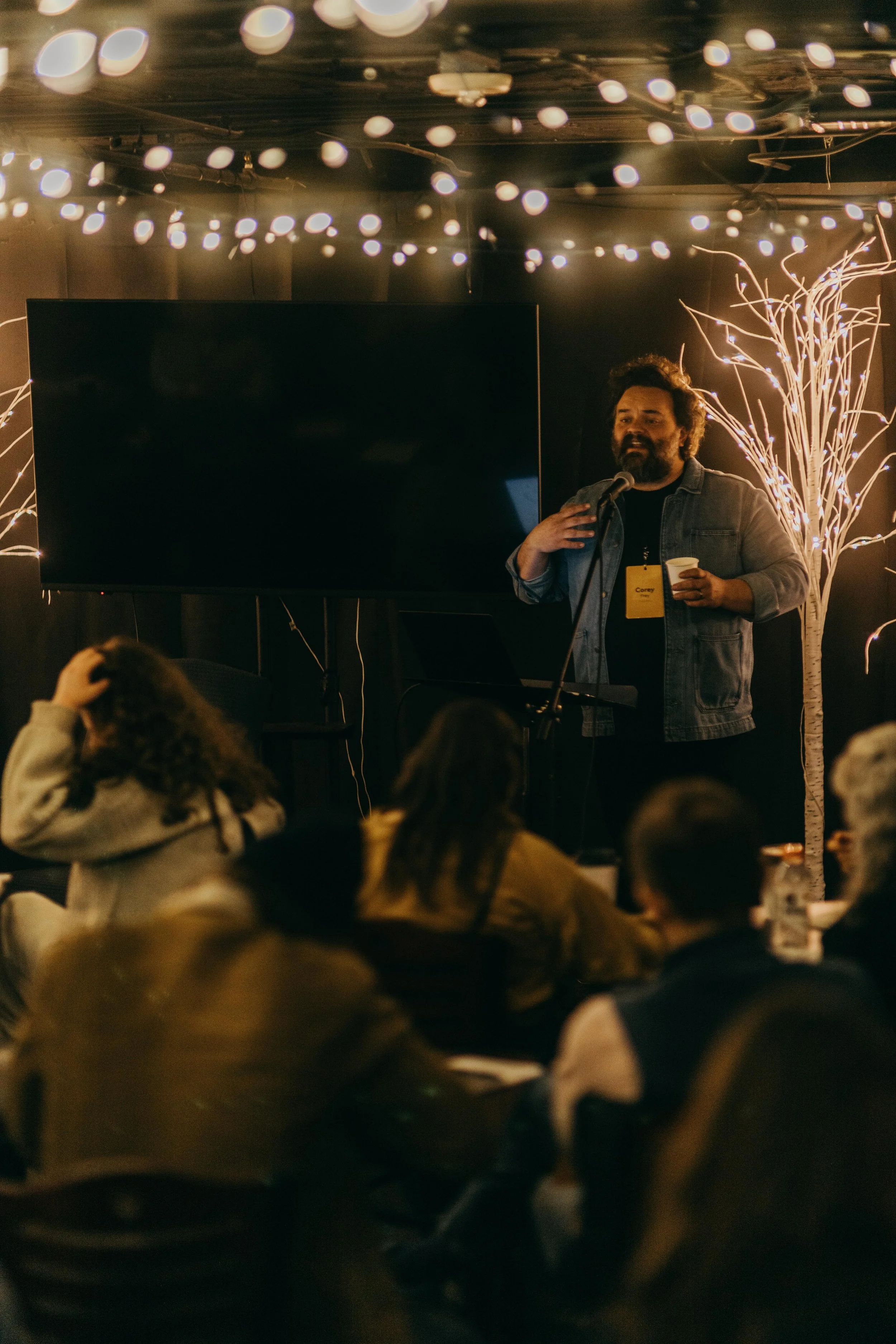 A man with dark curly hair and a beard is speaking into a microphone on a stage decorated with string lights and a white artificial tree. The audience is seated in front of him.