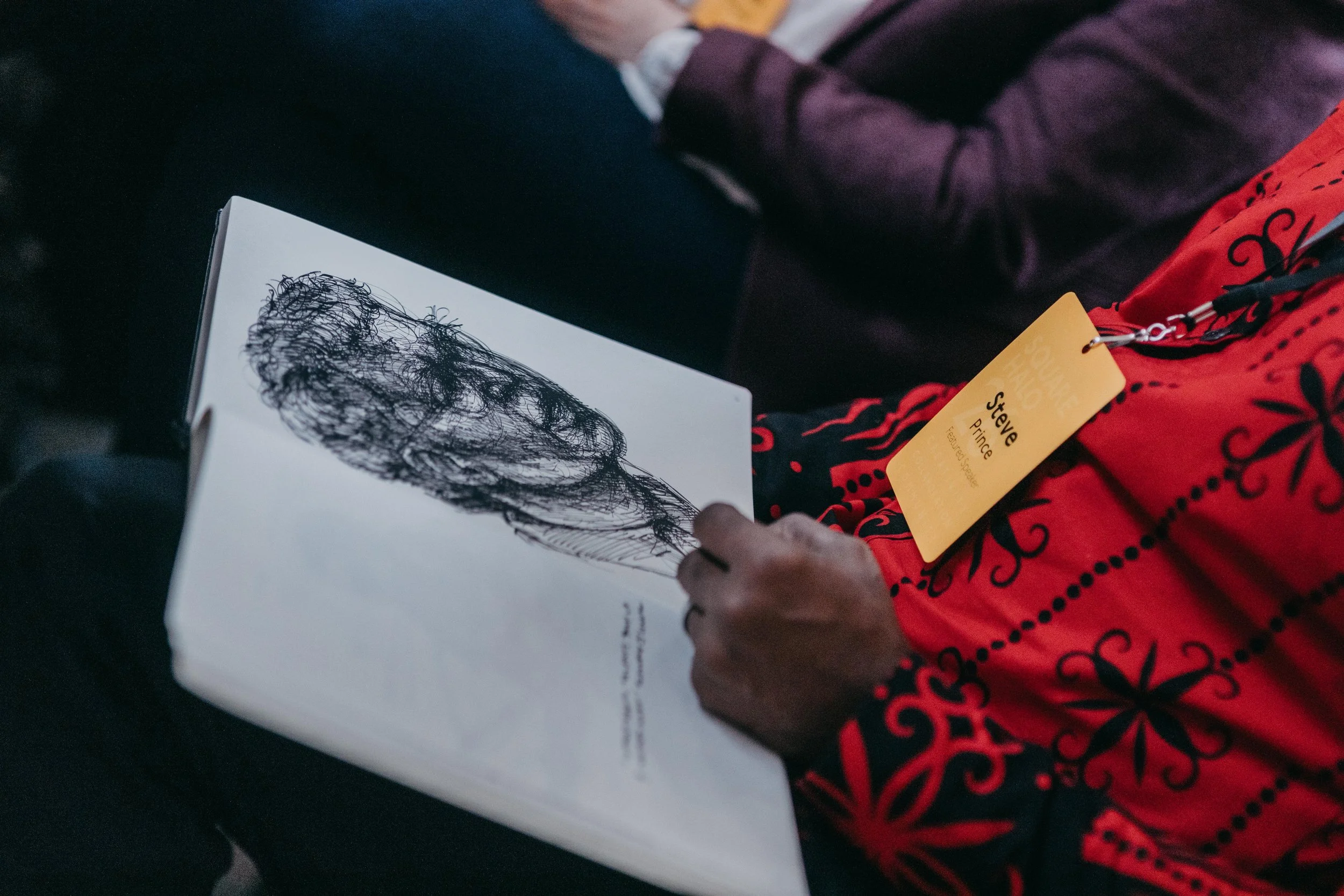 Person with dark skin and maroon shirt looking at a book with a black scribble drawing on the page.