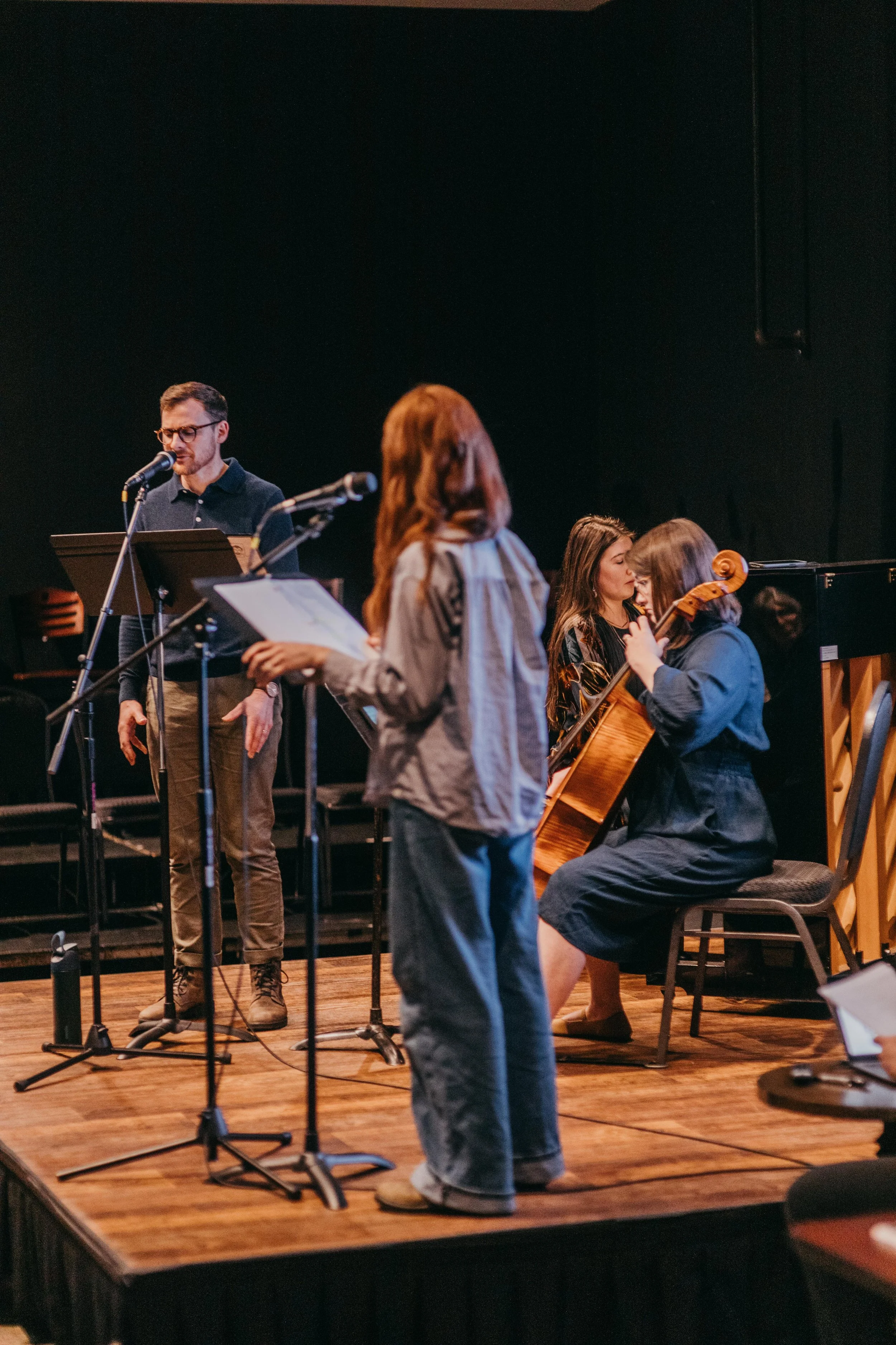 A group of five musicians preparing for a performance on a stage with a black background. One man is singing into a microphone and reading sheet music, while three women are engaged in different activities, one holding a cello. The stage has wooden f