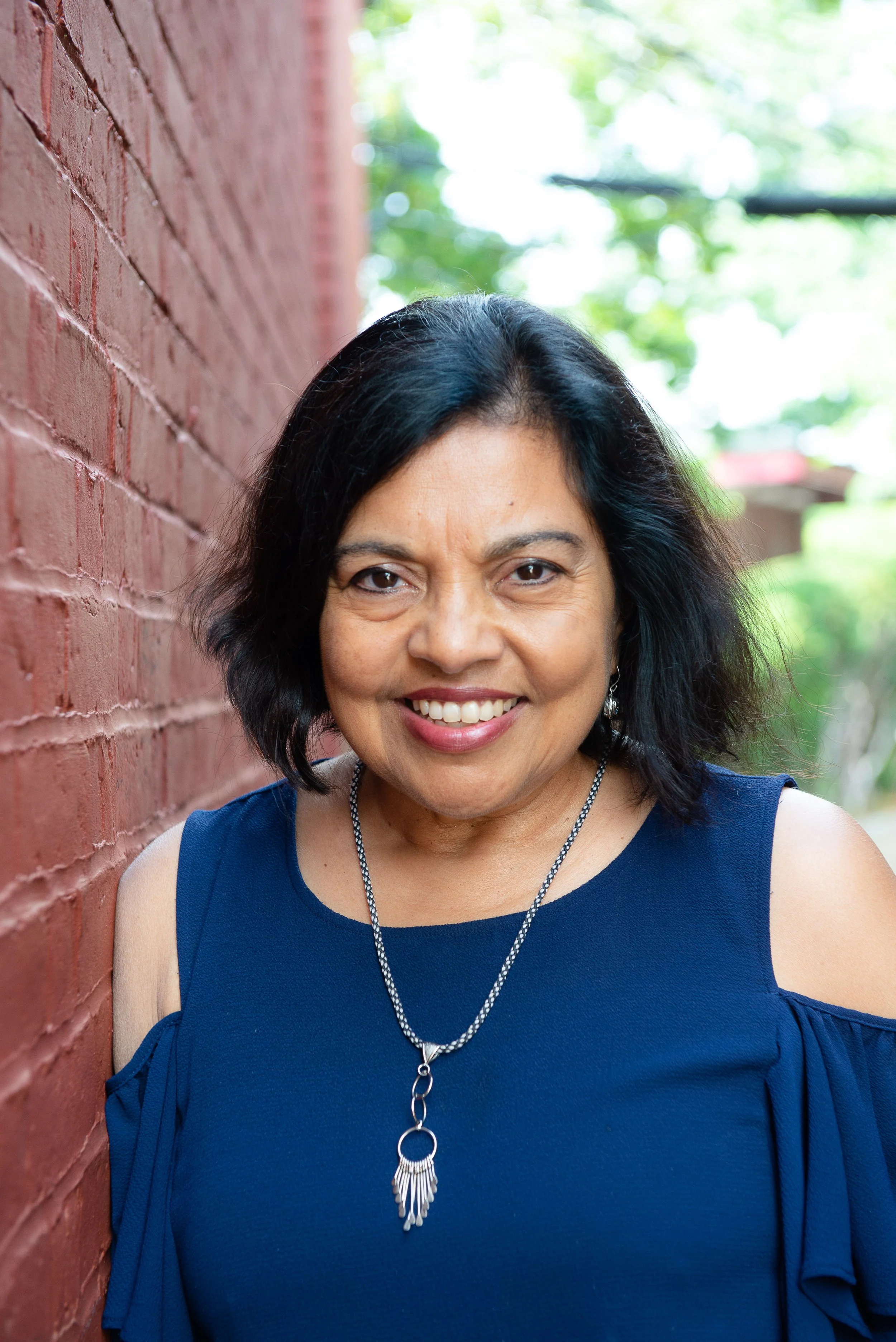 A middle-aged woman with black hair, smiling, wearing a blue sleeveless top, standing outdoors near a red brick wall, with green trees and a bright sky in the background.