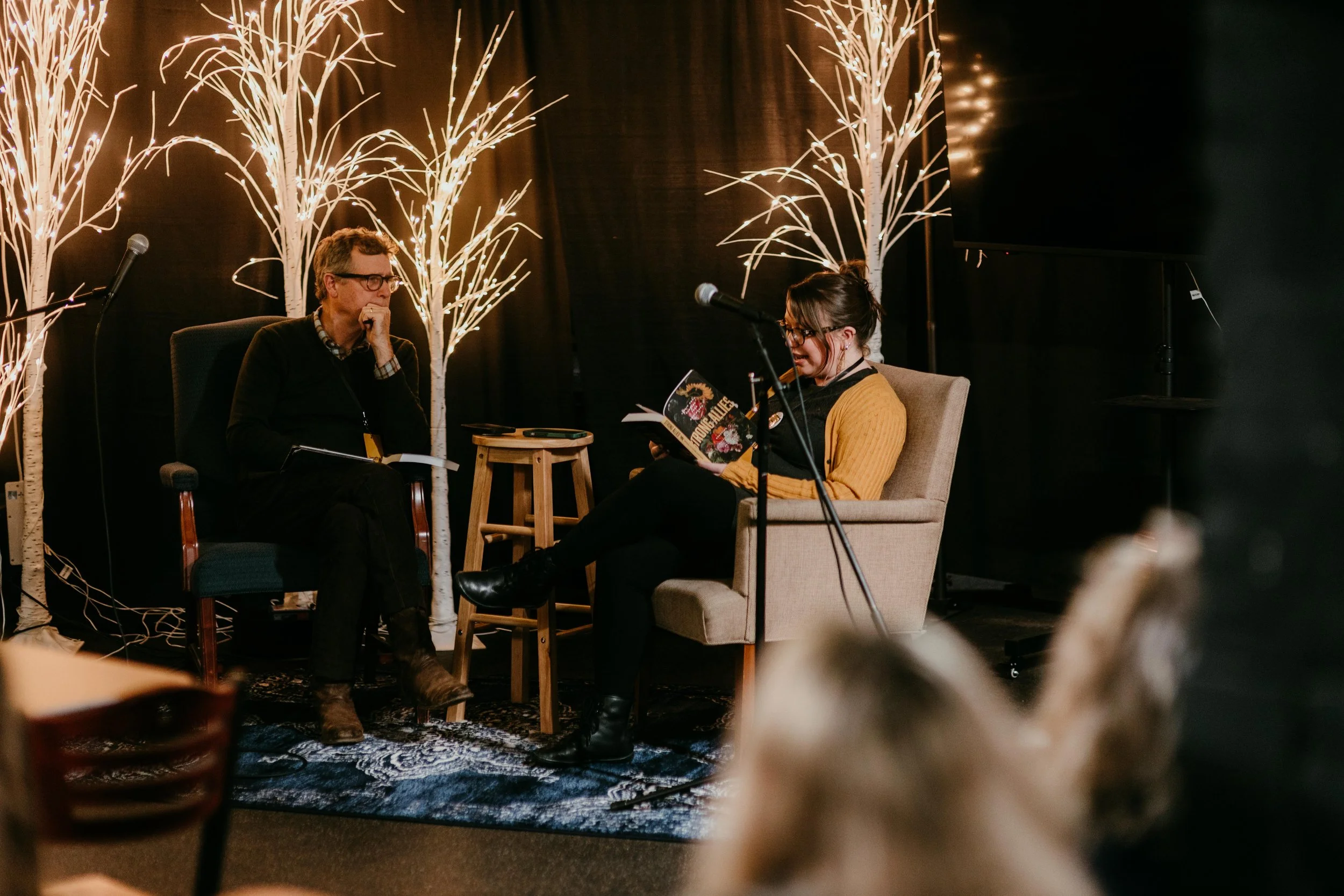 Two people seated on stage during an event, with decorative white light trees in the background. One person is reading from a booklet, the other is listening attentively.