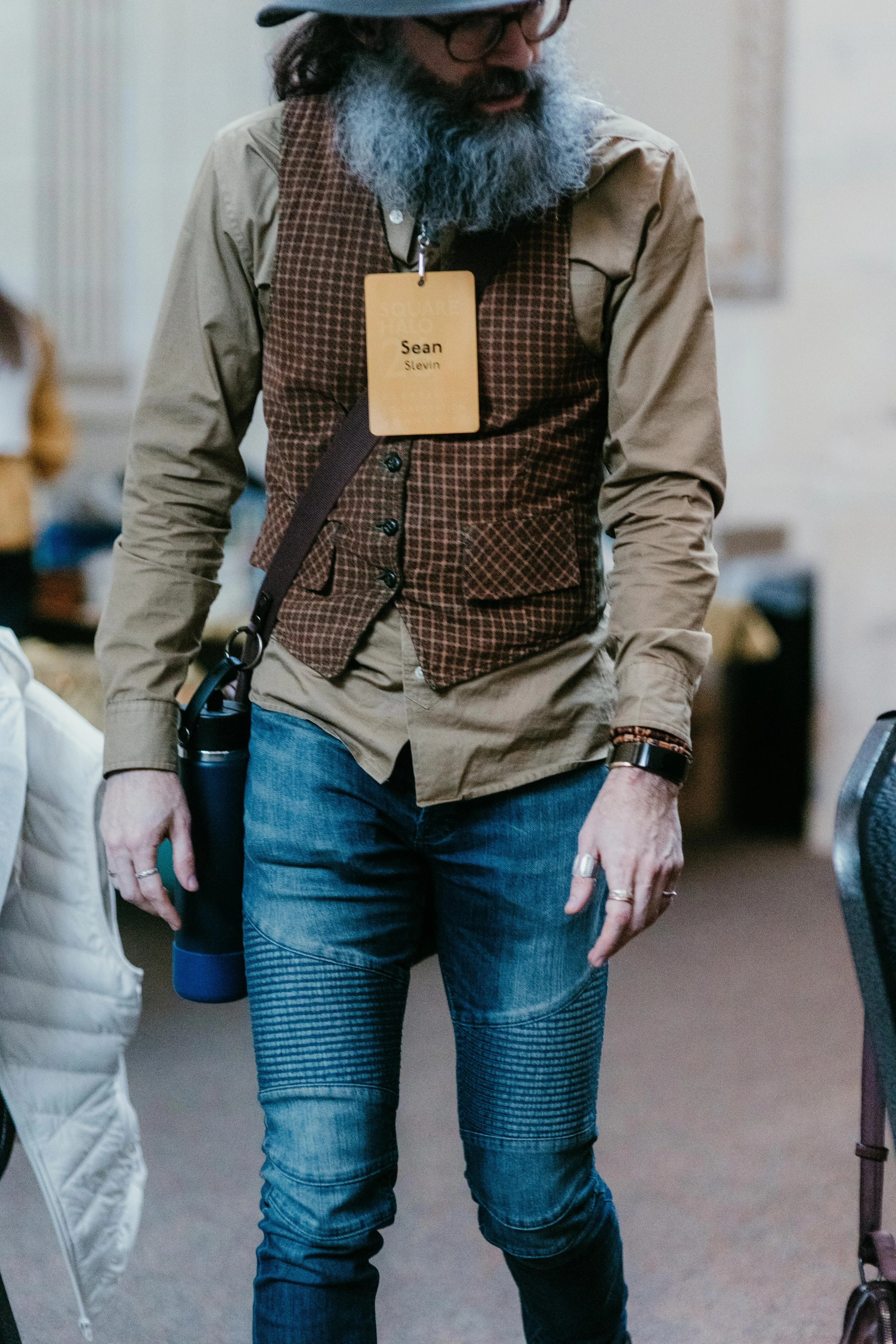 A person wearing a name badge labeled 'Sean Stevin' with a dark beard and glasses, dressed in a plaid vest over a beige shirt and blue jeans, standing indoors.