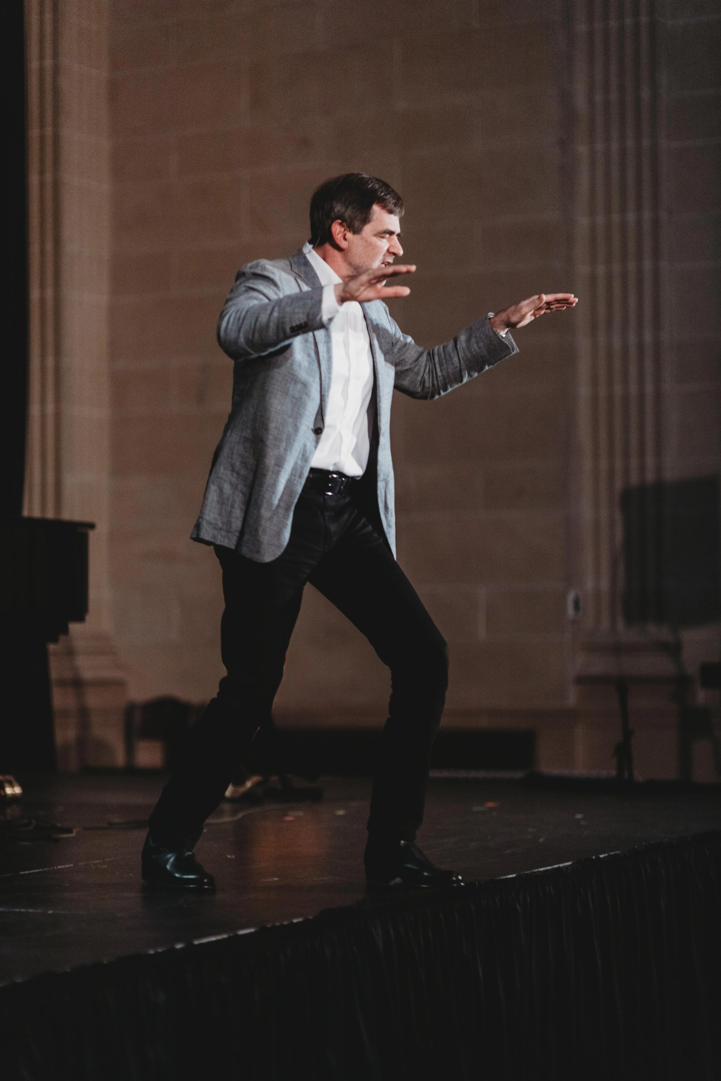 A man wearing a gray blazer, white shirt, and black pants is dancing on a stage with a dark background, with one hand raised and an intense expression on his face.
