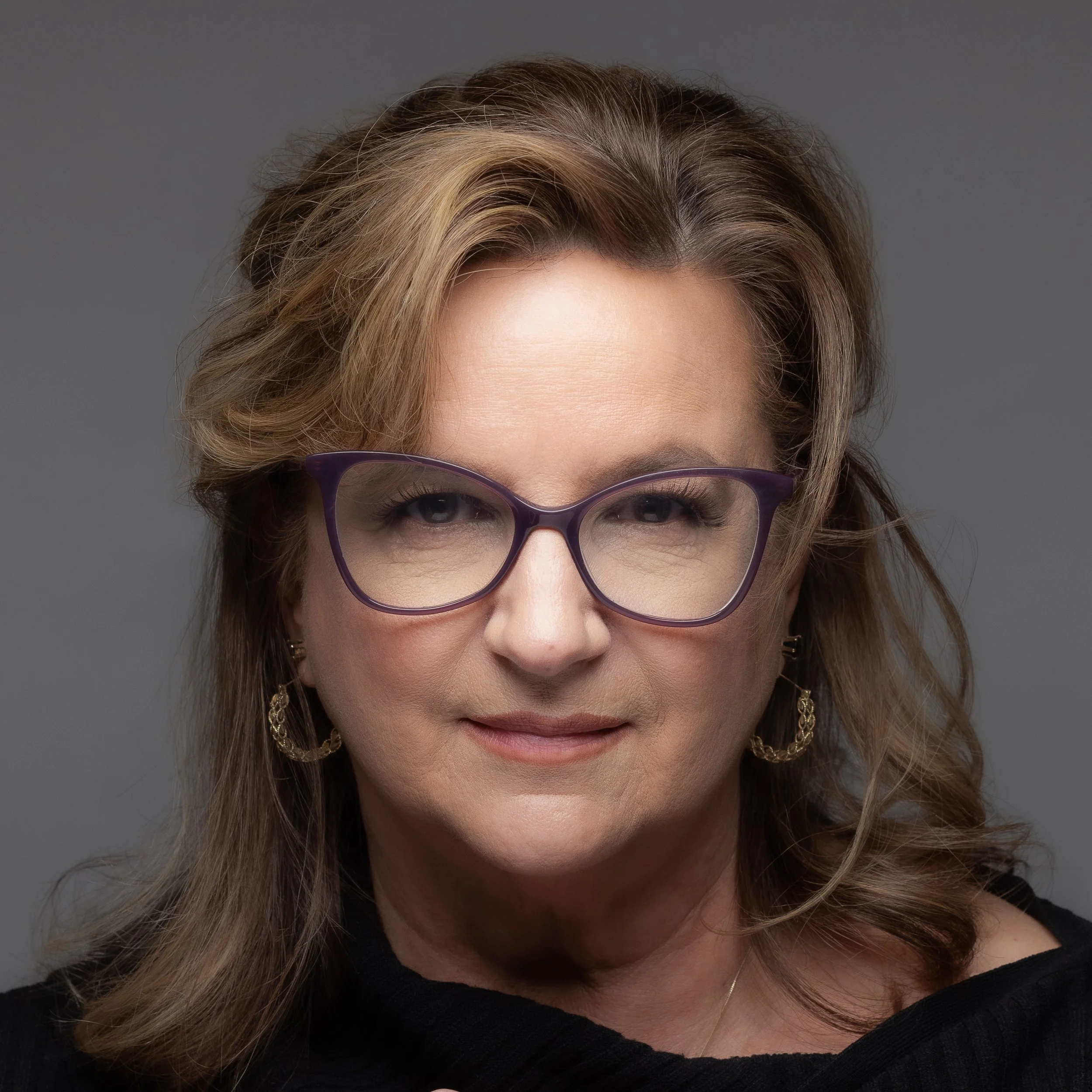 A woman with blonde, shoulder-length hair and glasses, wearing a black top and gold earrings, against a grey background.