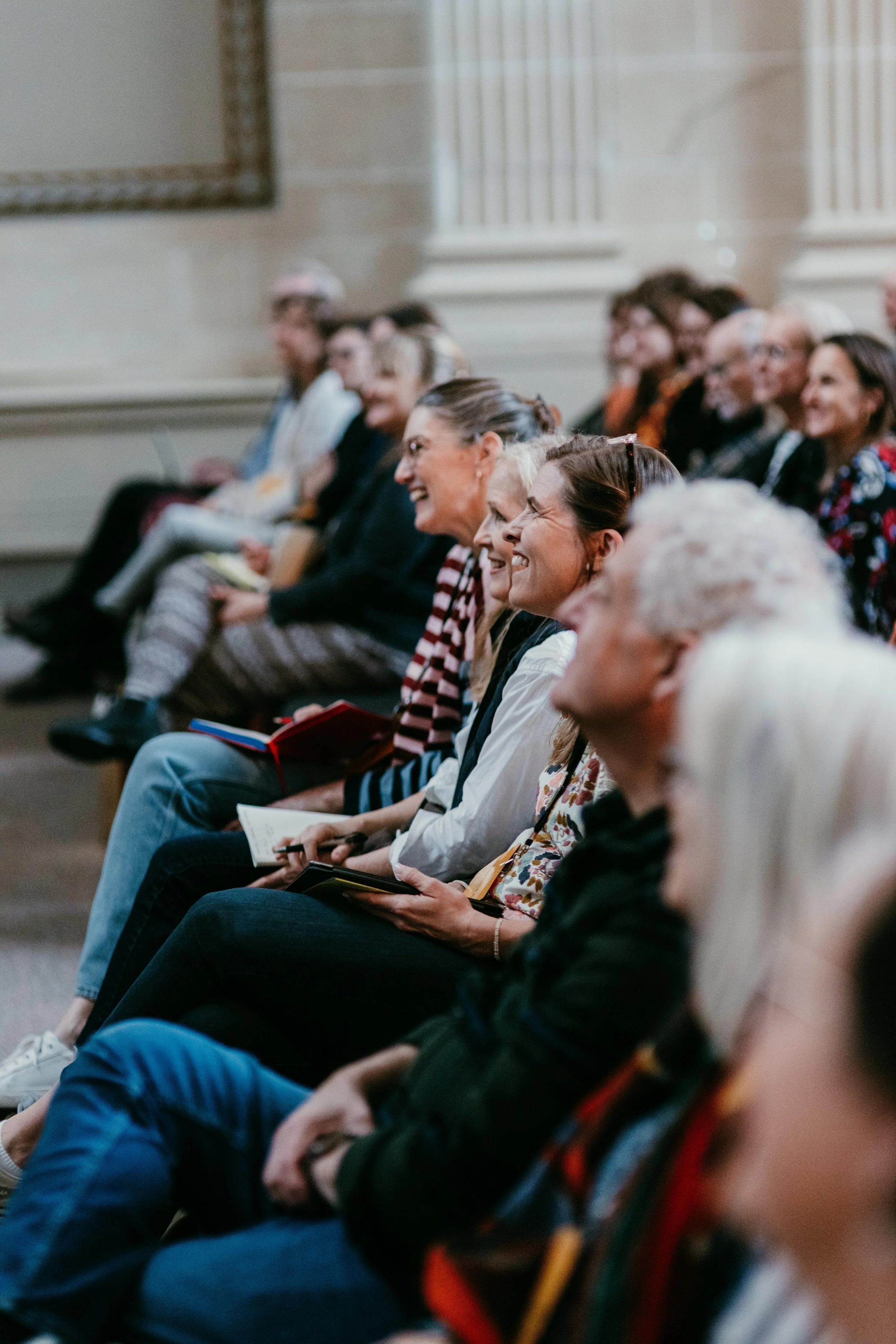 Group of people sitting in an audience, laughing and holding notebooks or papers.