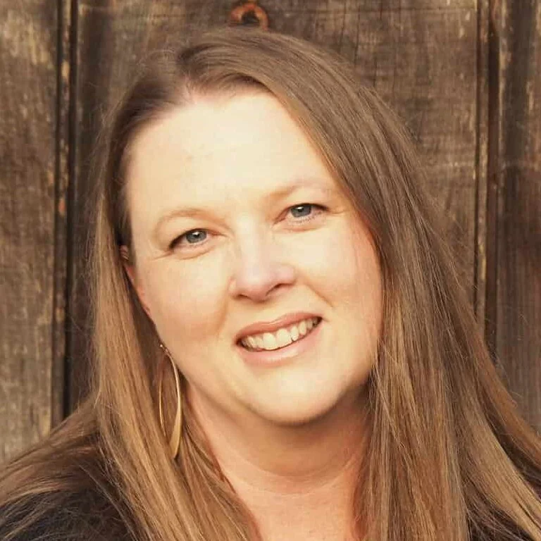 A woman with long brown hair, blue eyes, and fair skin smiling in front of a wooden background.