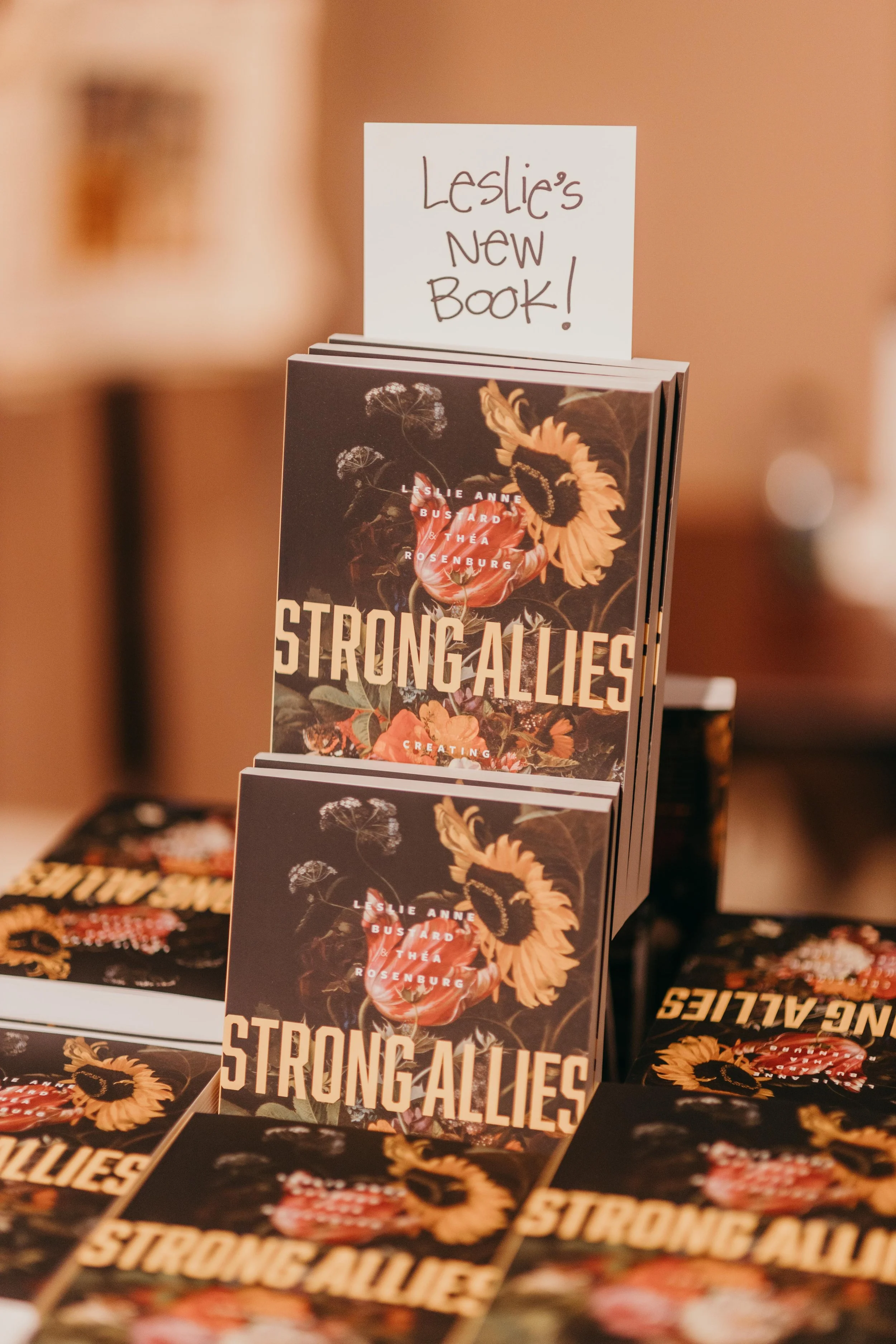 A display stand featuring a book titled 'Strong Allies' by Leslie Anne Bustard and Thia Rosenburg, with a handwritten sign that says 'Leslie's new book!'