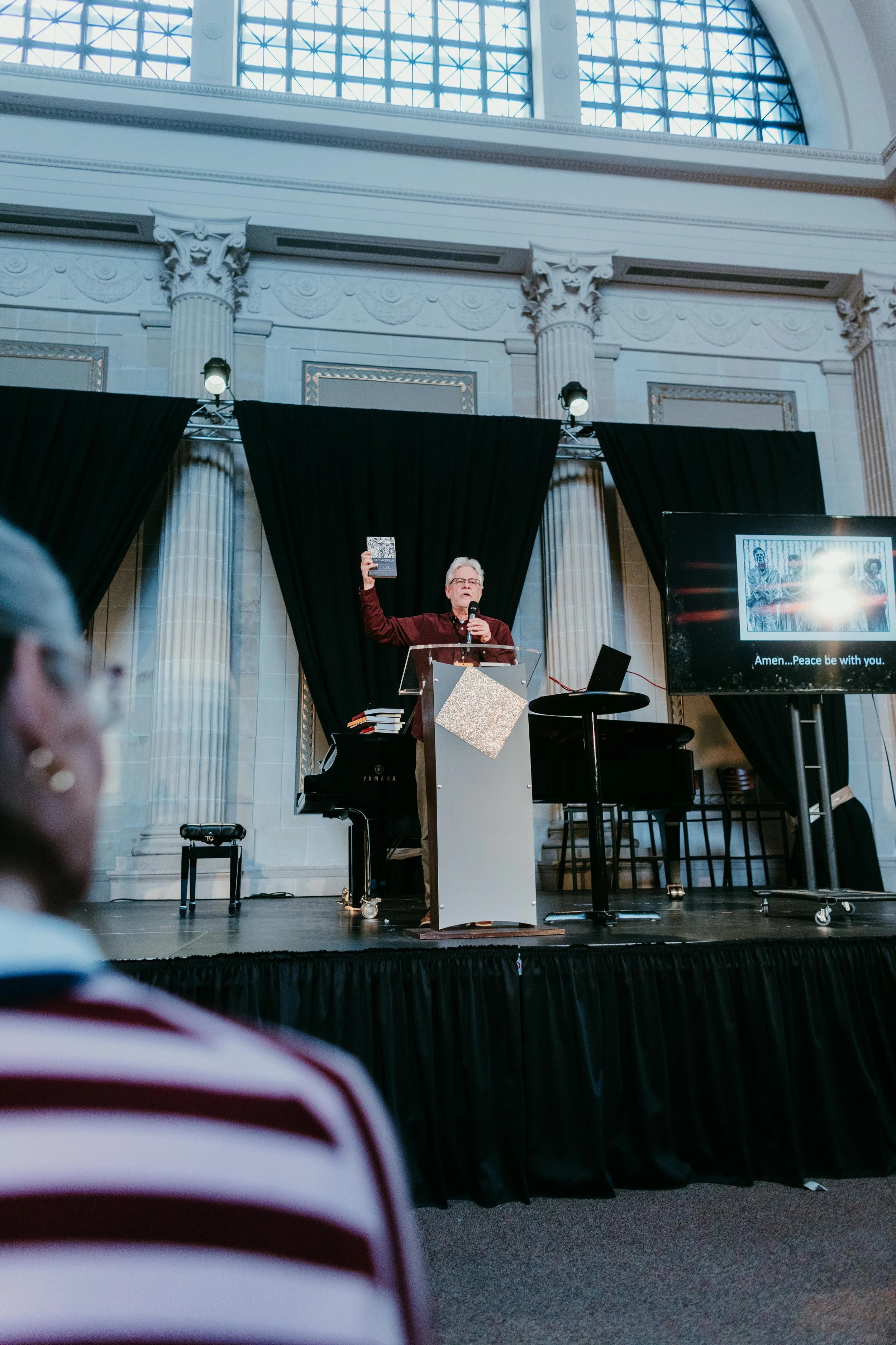 A man is speaking at a podium in a large hall with tall columns and a high arched window ceiling. He is holding a booklet in one hand and a microphone in the other. There is a piano nearby and a large screen displaying a group of people and the text 