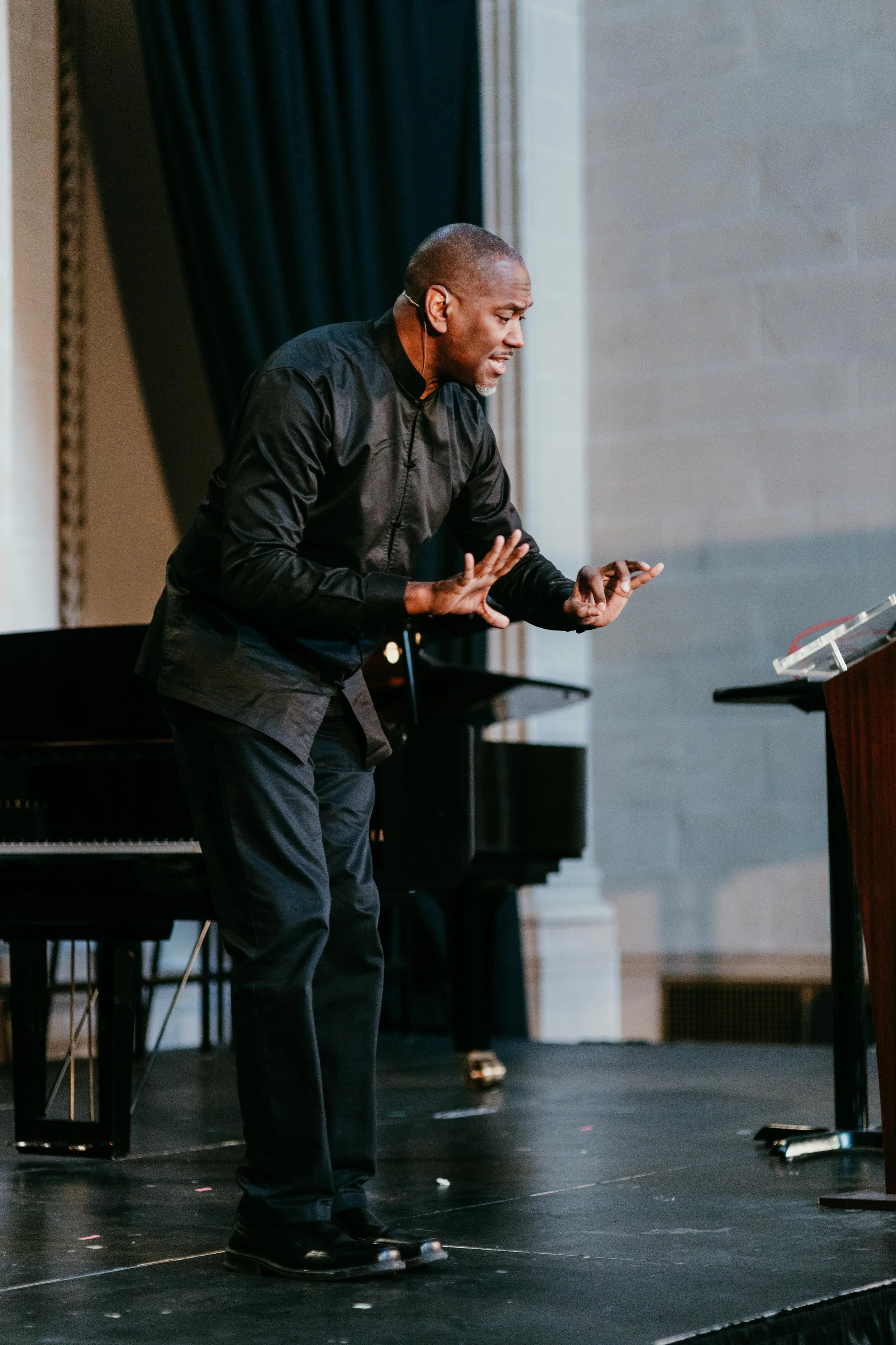 A man in black clothing performing on stage, possibly speaking or singing, with a grand piano in the background and a music stand to the right.