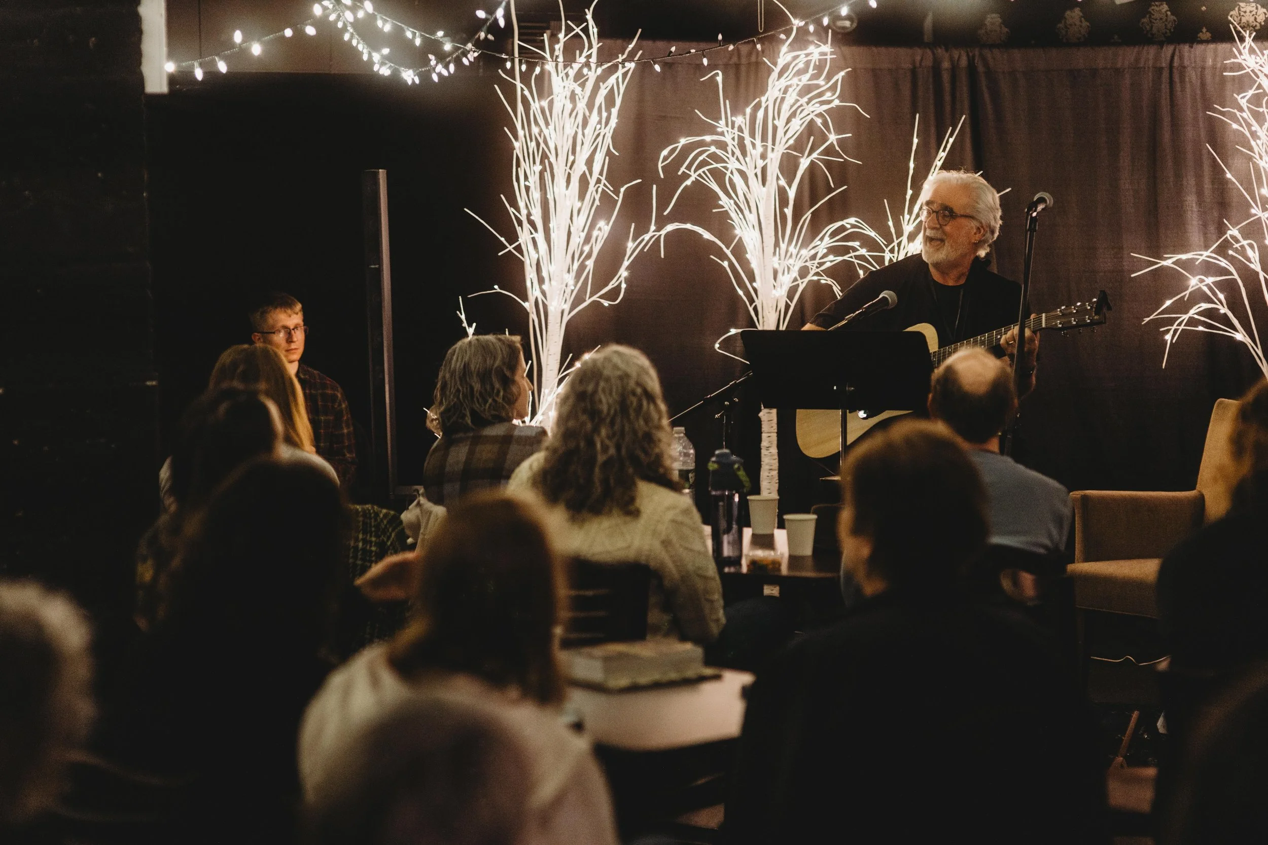 An older man with gray hair and glasses playing an acoustic guitar and singing into a microphone at a small indoor concert or open mic event. Audience members are seated and listening, with dark curtains and decorative string lights with white branch