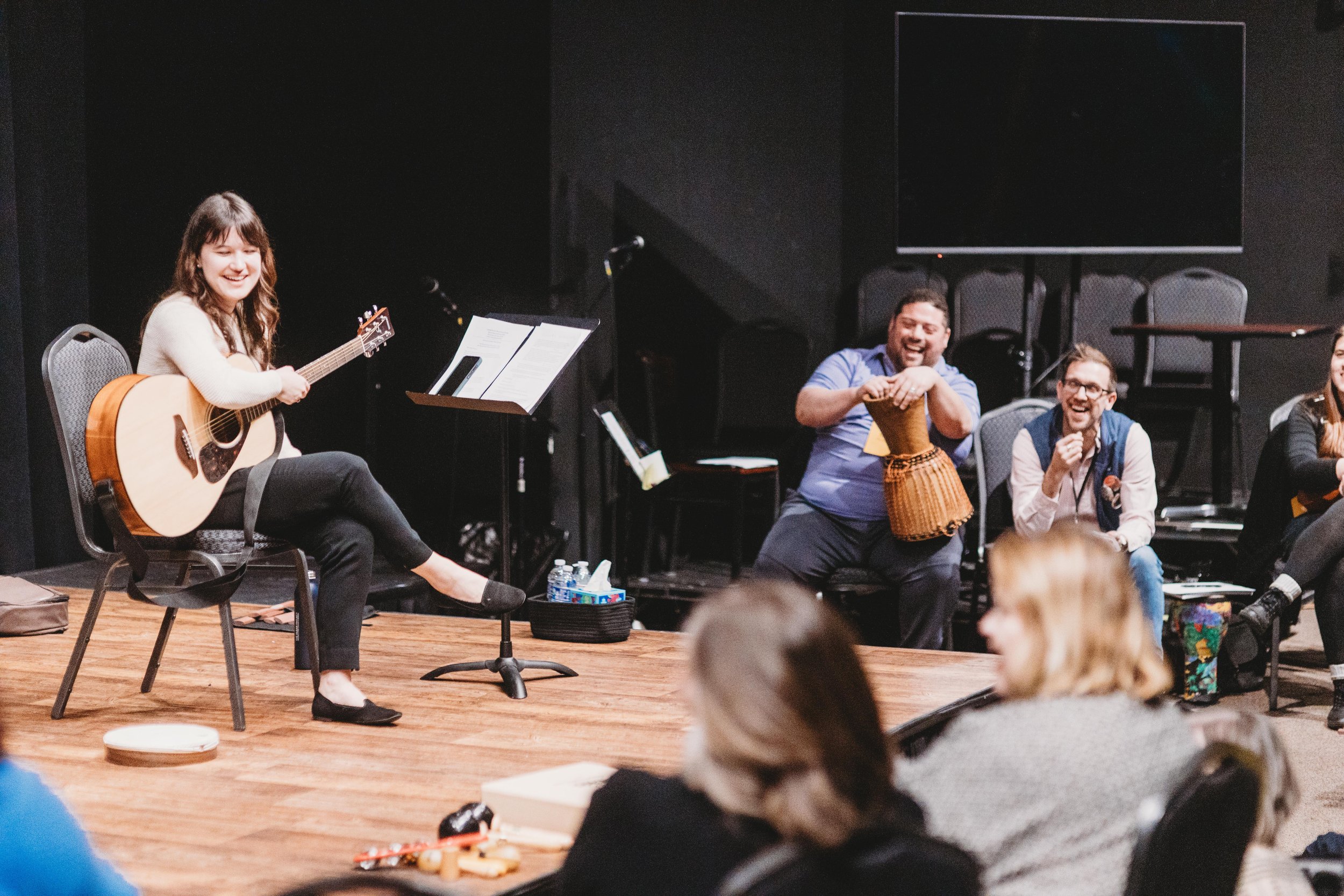 A woman playing an acoustic guitar on stage while people in the audience laugh and smile, with some sitting on the floor and others standing.