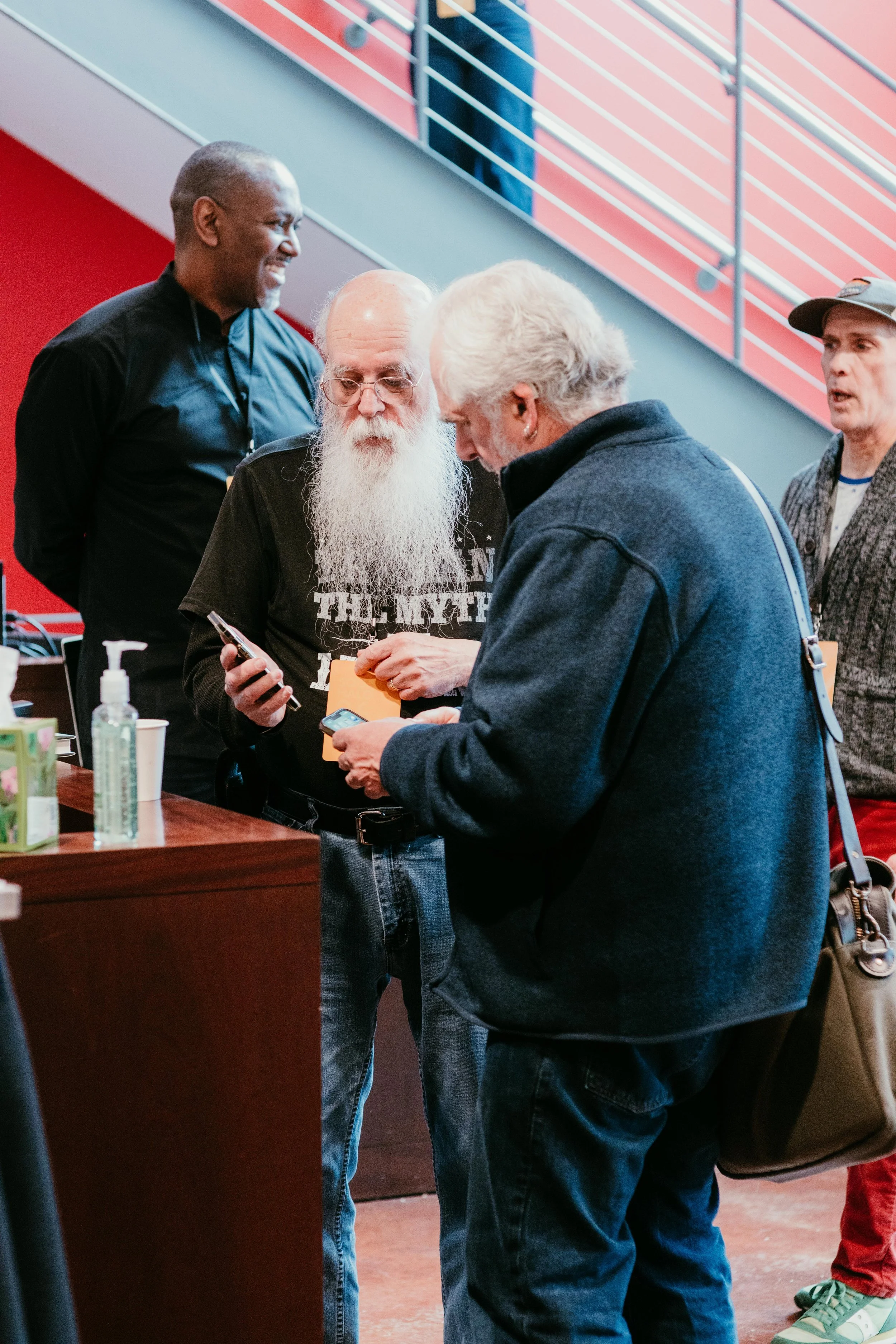 Four men are gathered around a table, looking at a phone. One man with a long white beard and glasses is holding the phone, others are looking at it with interest. The setting appears to be indoors, possibly at an event or conference.