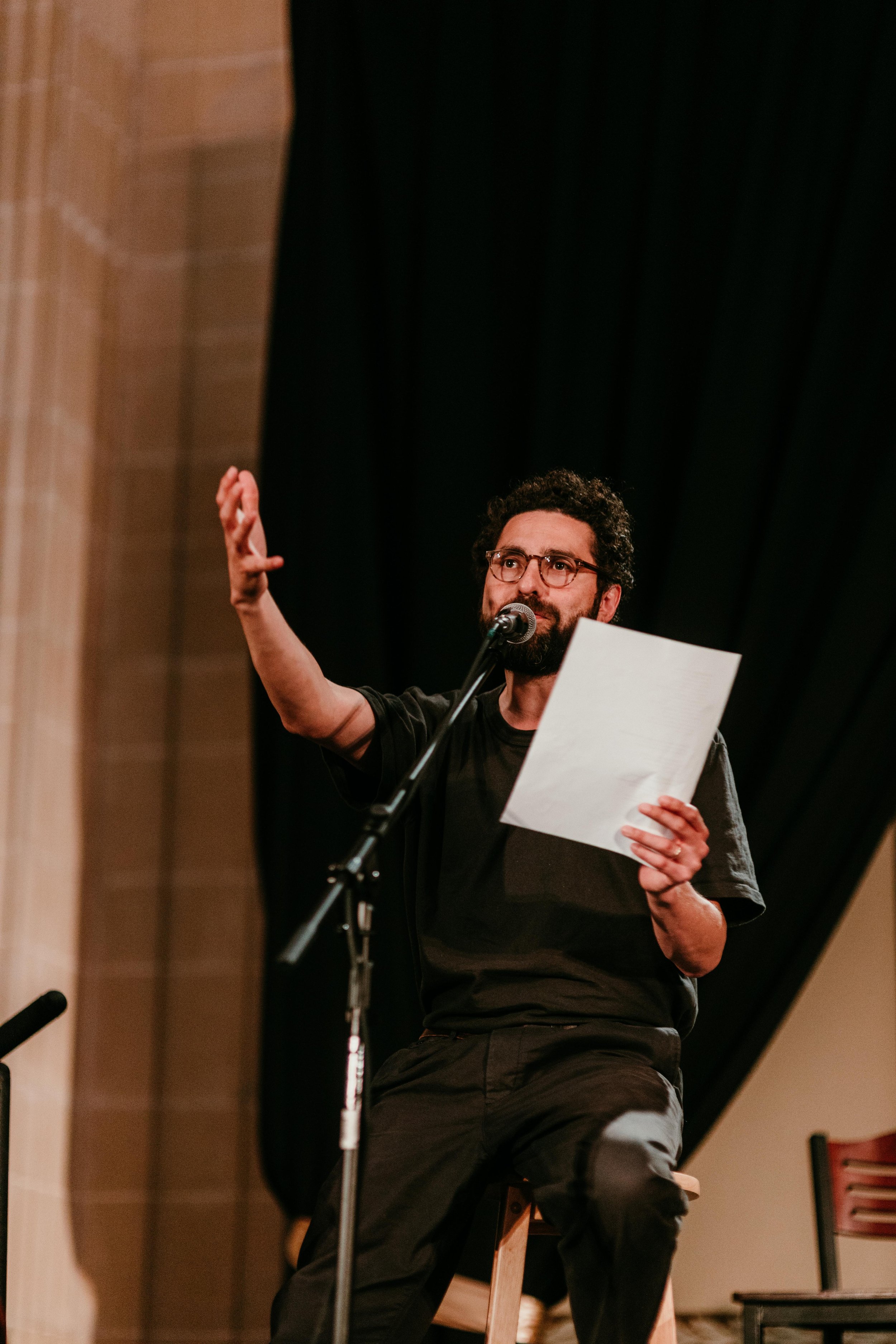A man with glasses and curly hair is speaking into a microphone while holding a paper. He is sitting on a stool on a stage with a dark curtain backdrop.