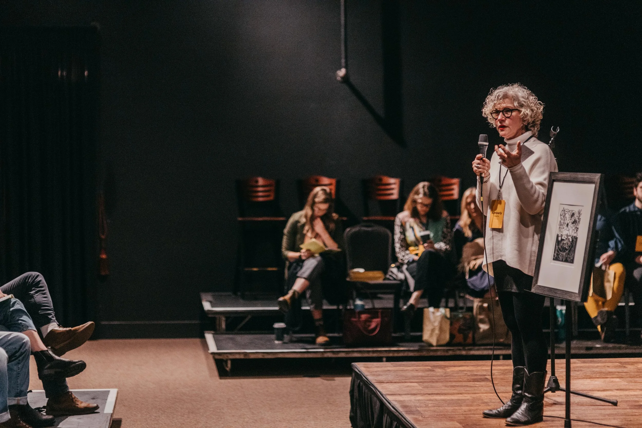 A woman with curly gray hair, glasses, and a white sweater speaking into a microphone on stage during an event, with a framed artwork displayed on an easel next to her, while other seated women are seen in the background.