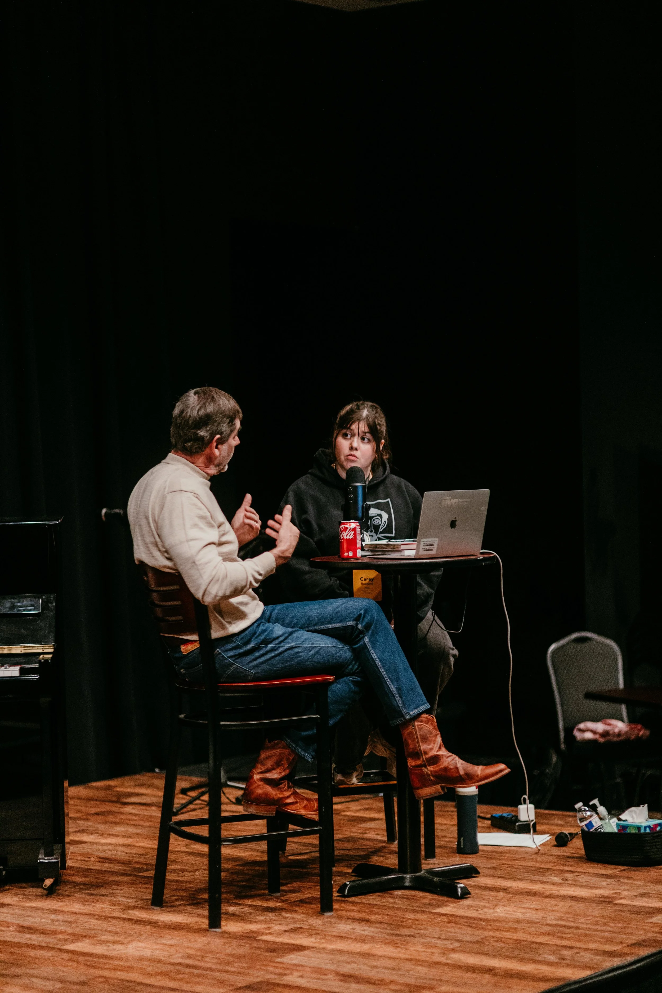 Two people are engaged in a conversation at a panel or discussion event; one person with gray hair and beige shirt is talking, while the young woman with dark hair and hoodie listens with a serious expression. They are seated at a table with a laptop