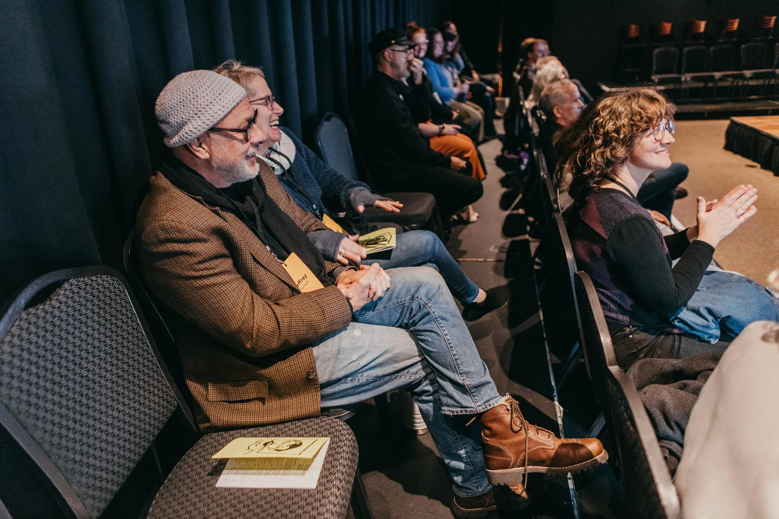People seated in a theater or auditorium, smiling and clapping during a presentation or performance.