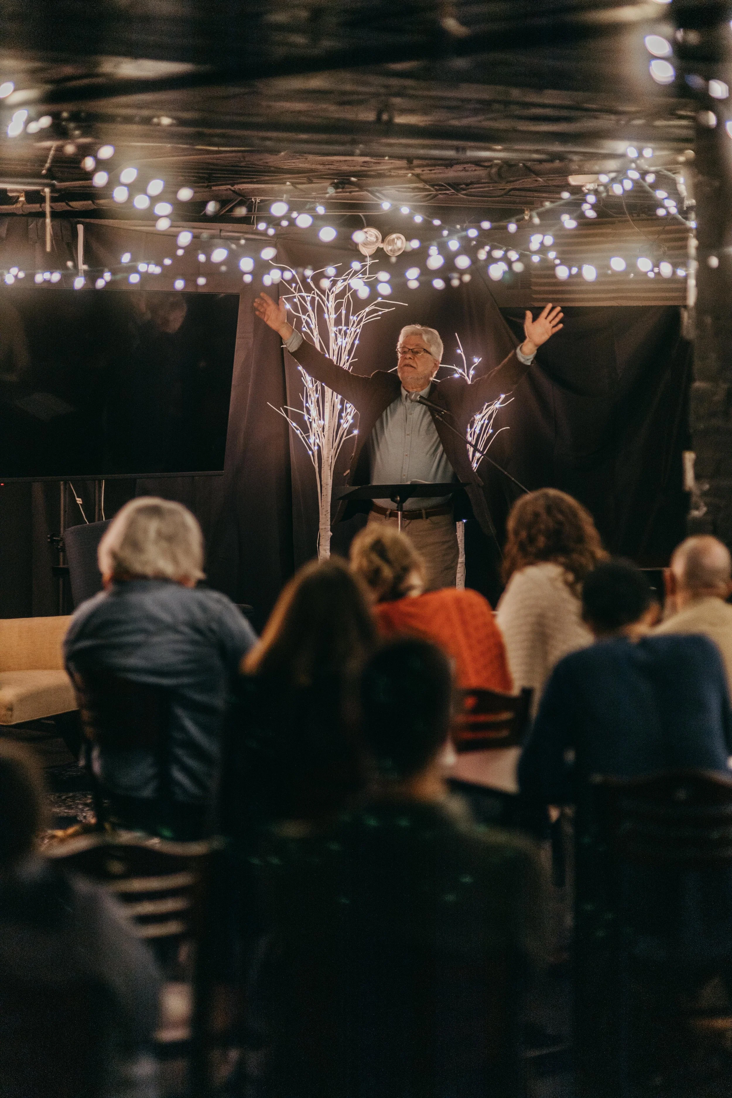 An elderly man with glasses is giving a speech or performance with his arms spread wide, behind a black curtain with decorative white branches. The room is decorated with string lights overhead and has an audience seated in front of him.