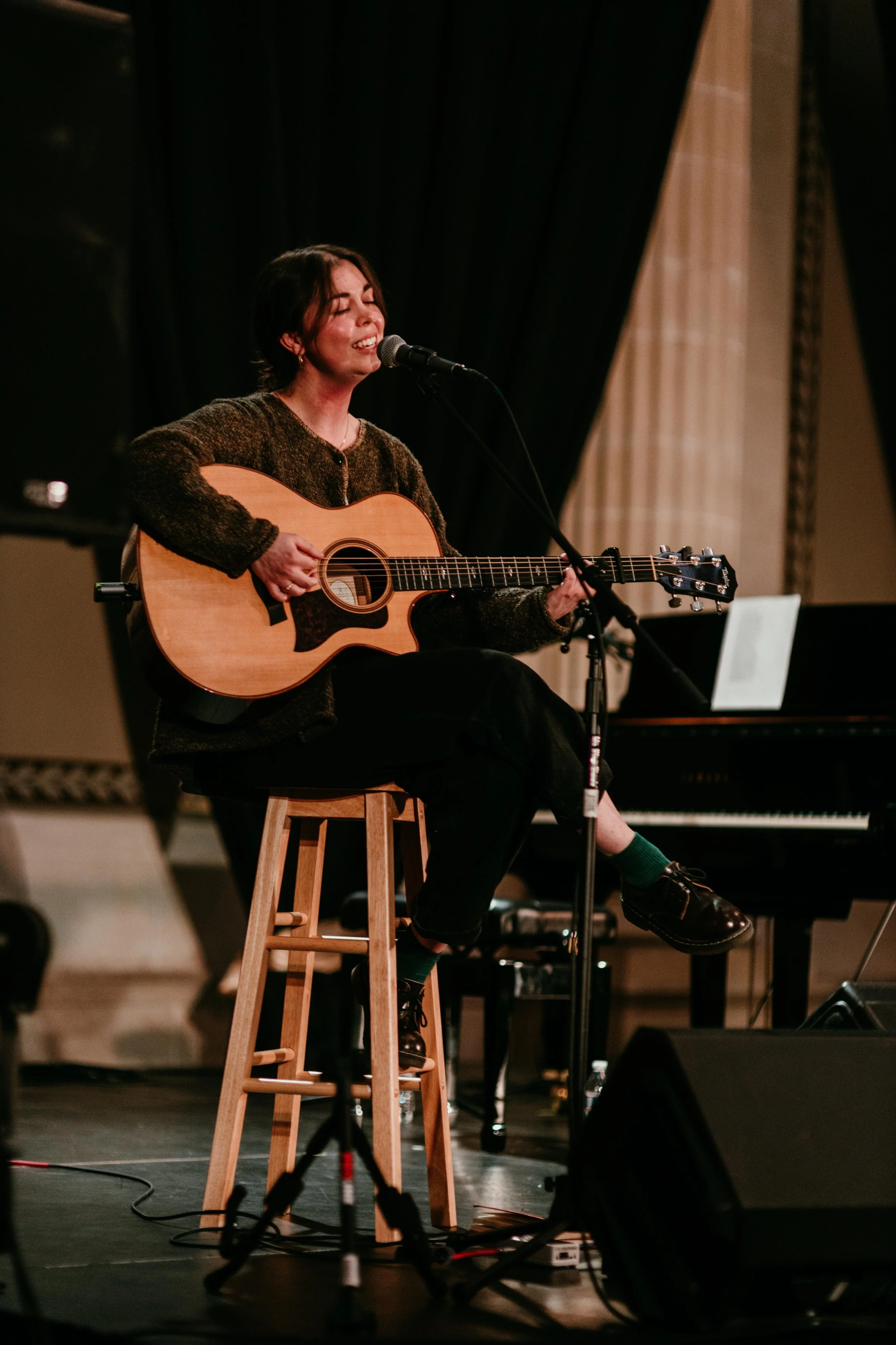 A woman sitting on a wooden stool playing an acoustic guitar and singing into a microphone on a stage with a black curtain in the background.