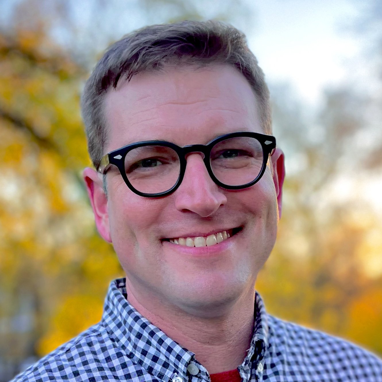 A smiling man wearing black-rimmed glasses and a checkered shirt outdoors during autumn with yellow and orange leaves in the background.