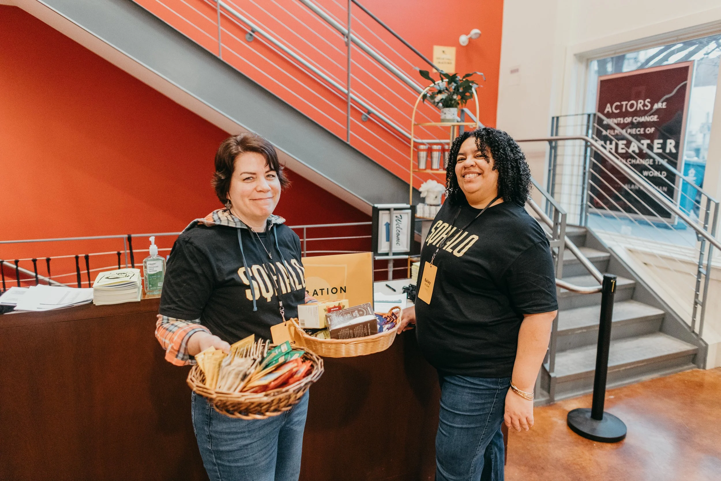 Two women standing at a reception desk, holding baskets of snacks and candies. The woman on the left has short brown hair, is wearing a black shirt with the word 'SOLANO' printed on it, and a hoodie layered underneath. The woman on the right has curl