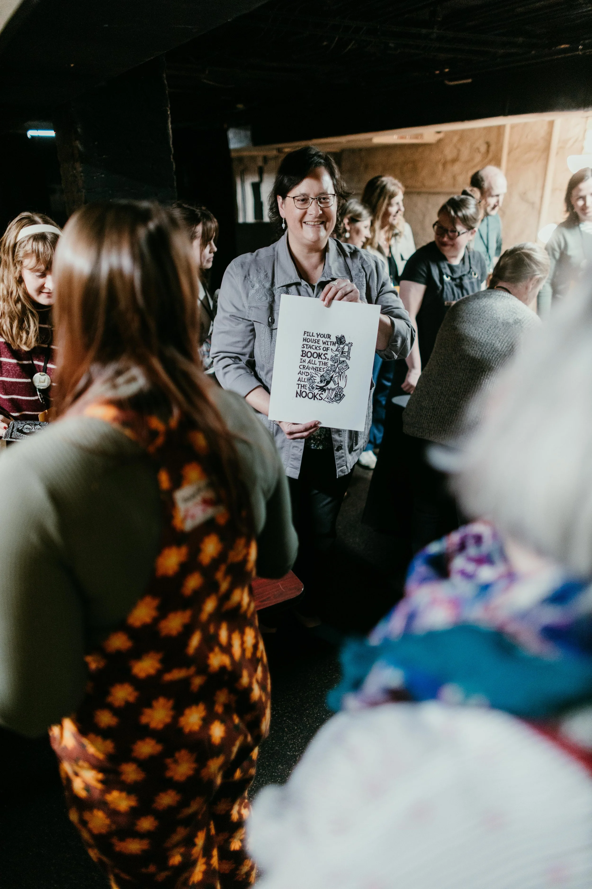 A group of people gathered indoors, with a woman in the center holding a sign that says, 'Fill your house with stacks of books, in all the crannies and all the nooks.' The room has wooden walls and warm lighting, and the people appear to be engaged i