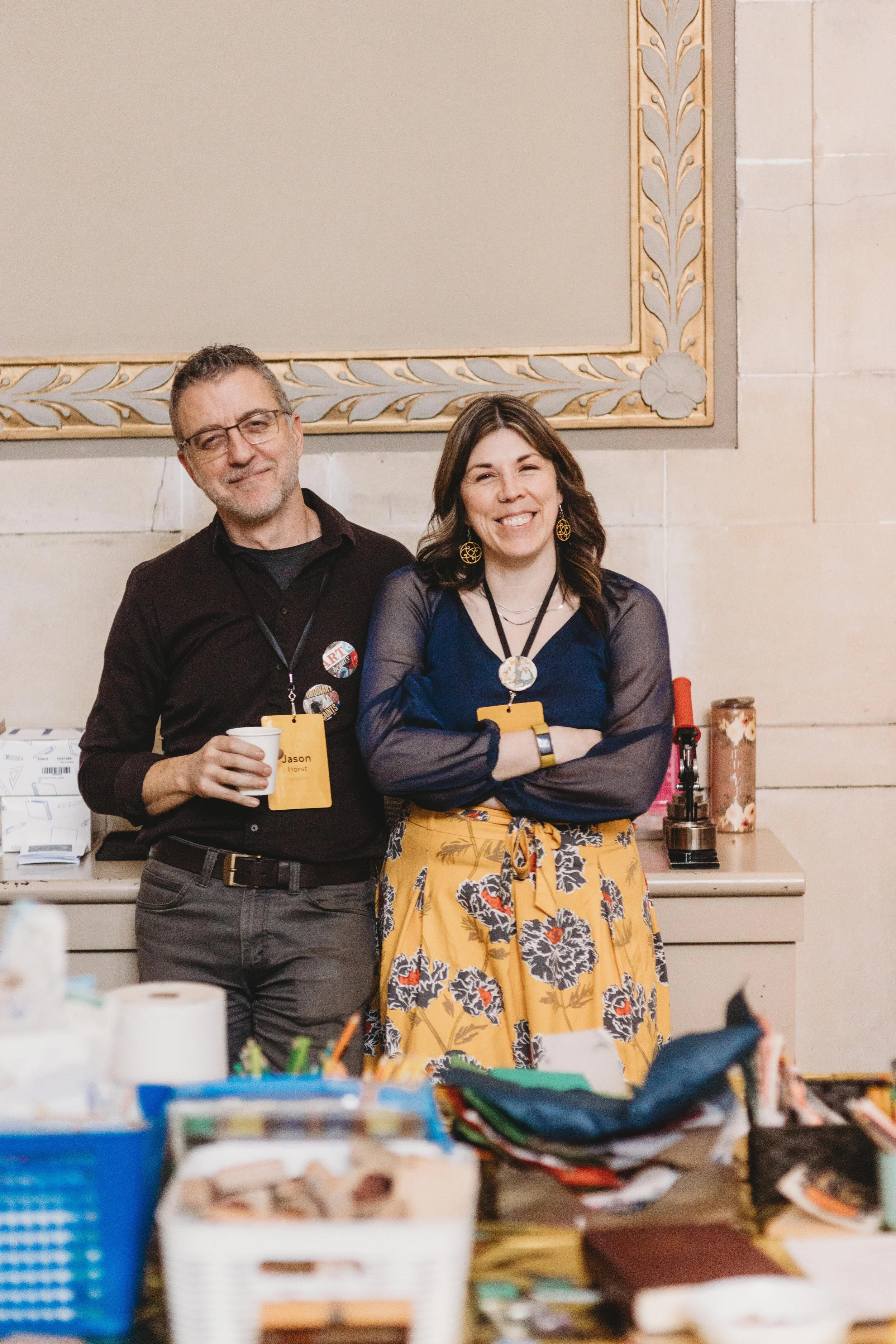 Two people standing together at a social event, smiling at the camera. One man is wearing glasses, a black shirt, and holding a white disposable cup. The woman is wearing a blue top, a yellow floral skirt, large earrings, and a badge around her neck.