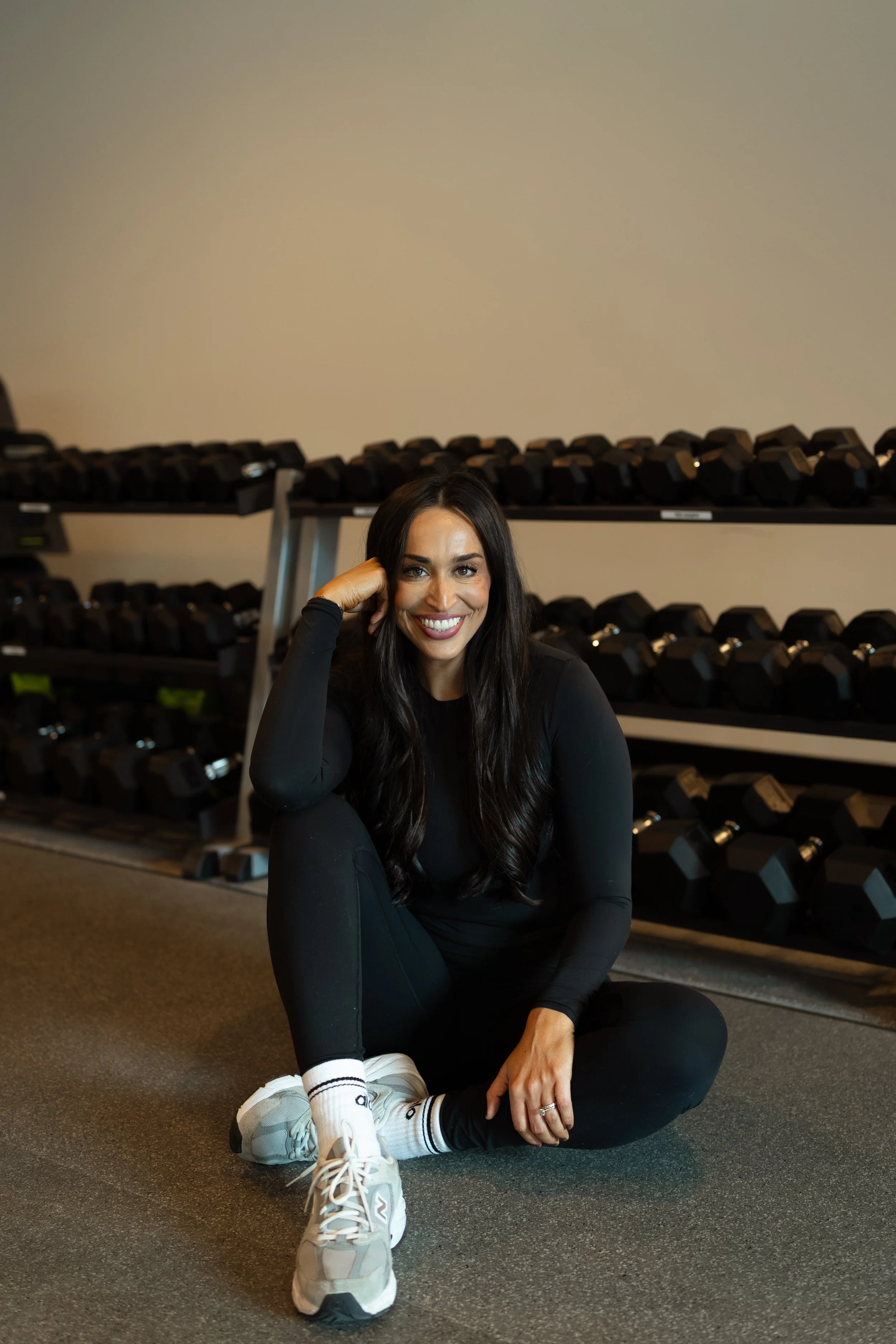 A smiling woman with long dark hair, wearing a black sleeveless top, poses with her arms crossed in a room with blue lighting.
