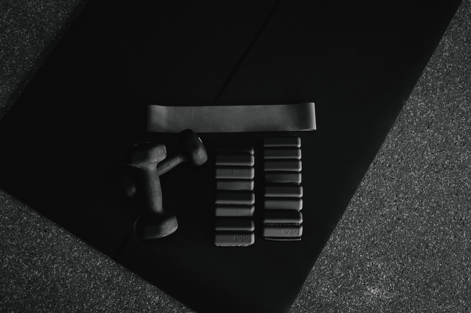 A black massage gun, a hammer, and six gray workout plates arranged on a dark surface in a minimalistic setting.